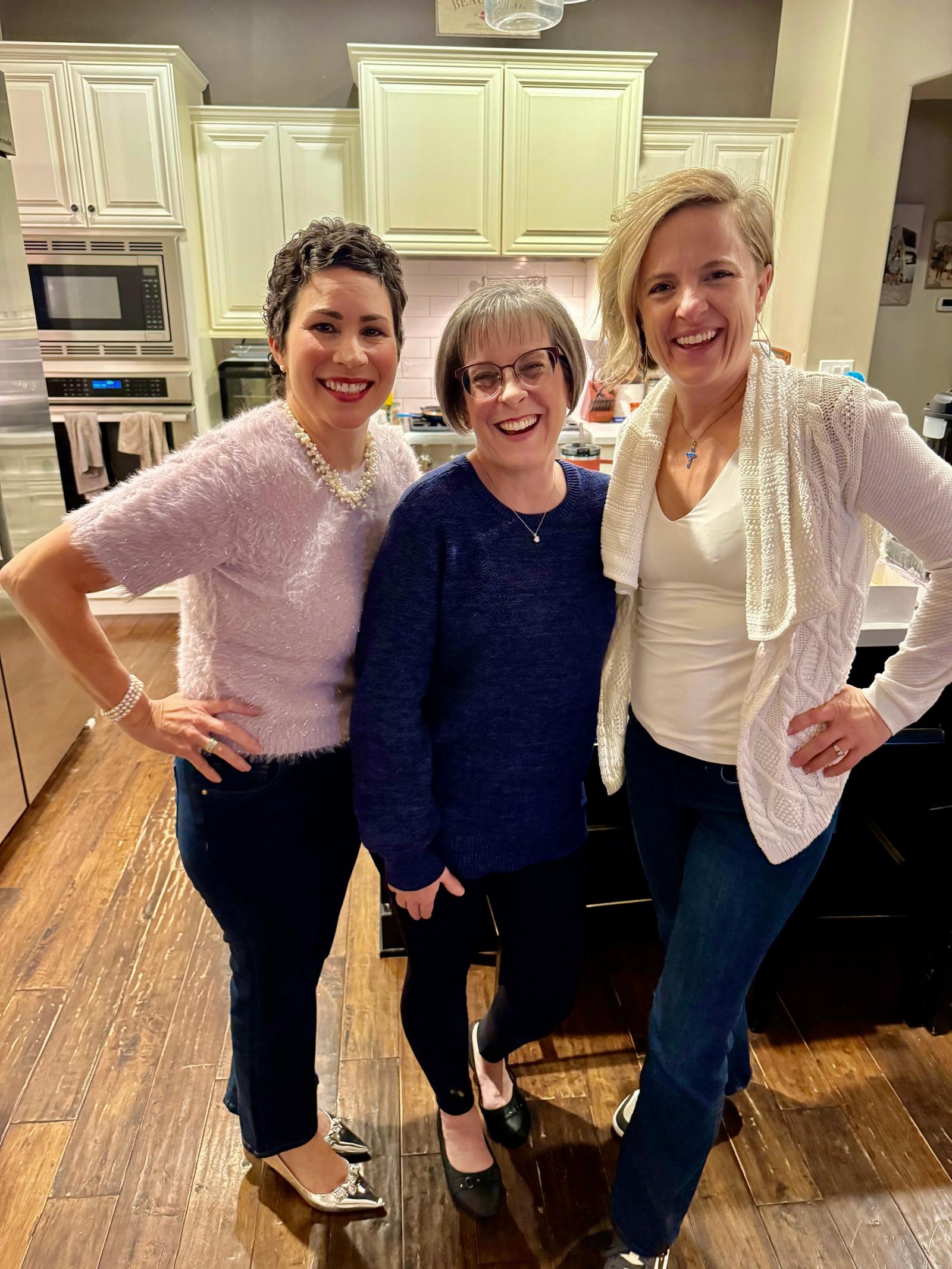 Three women smiling together in a kitchen at a 2025 Christmas party.