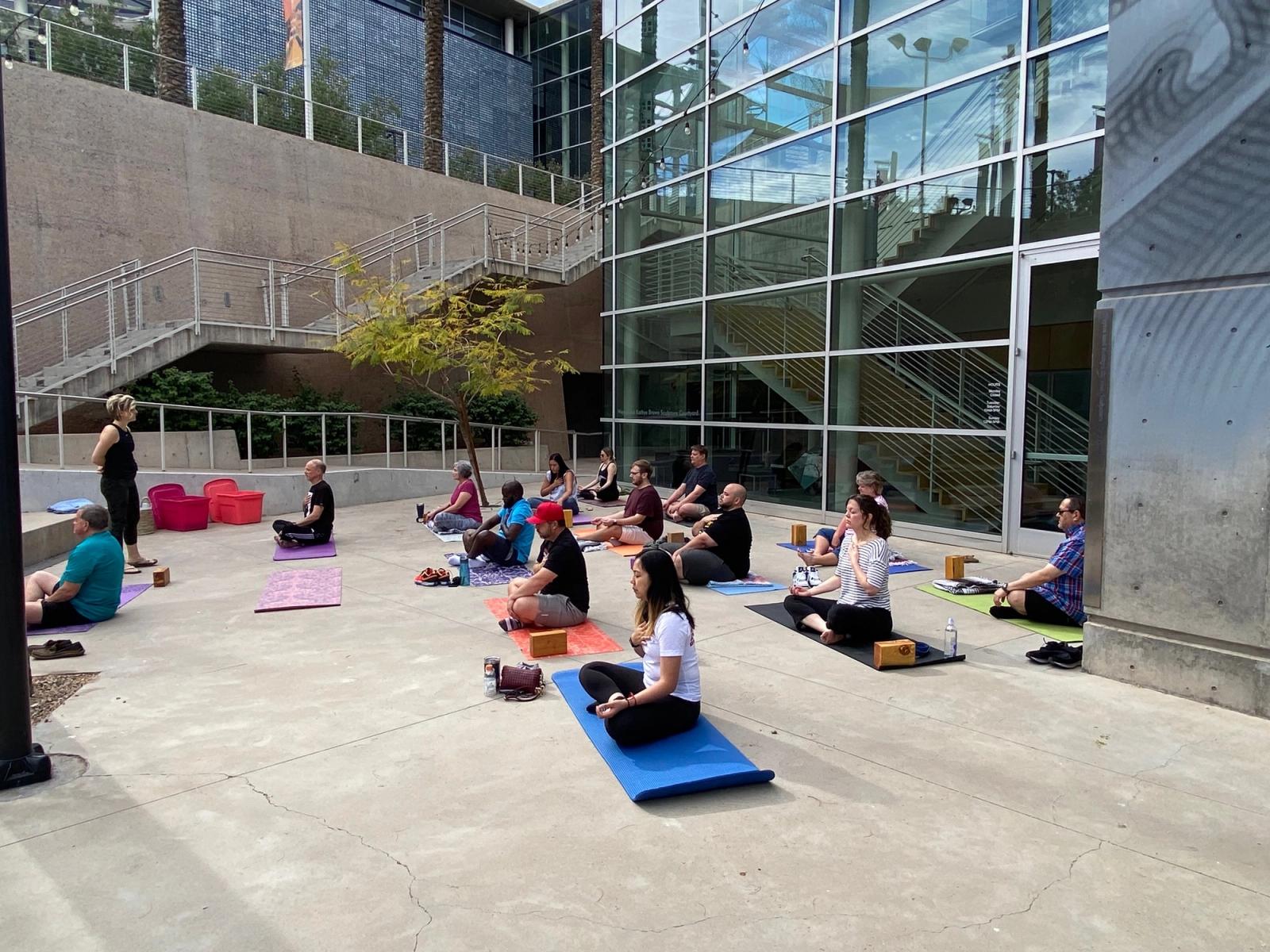 Participants seated on yoga mats outdoors beside a glass office building during a workplace mindfulness session.
