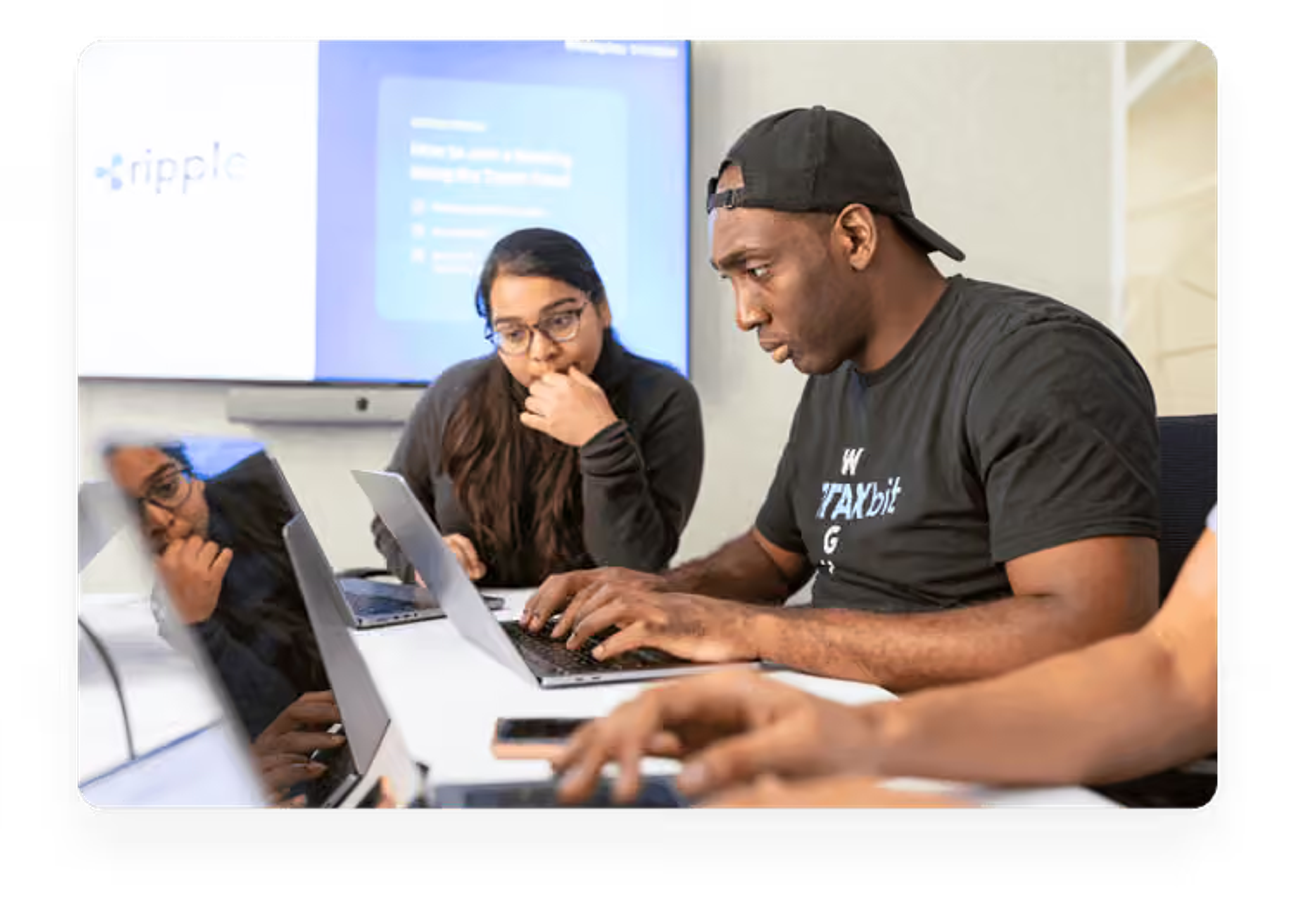 Two colleagues collaborating over laptops at a white table in a bright office environment, with a presentation screen visible in the background