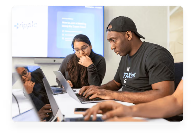 Two colleagues collaborating over laptops at a white table in a bright office environment, with a presentation screen visible in the background