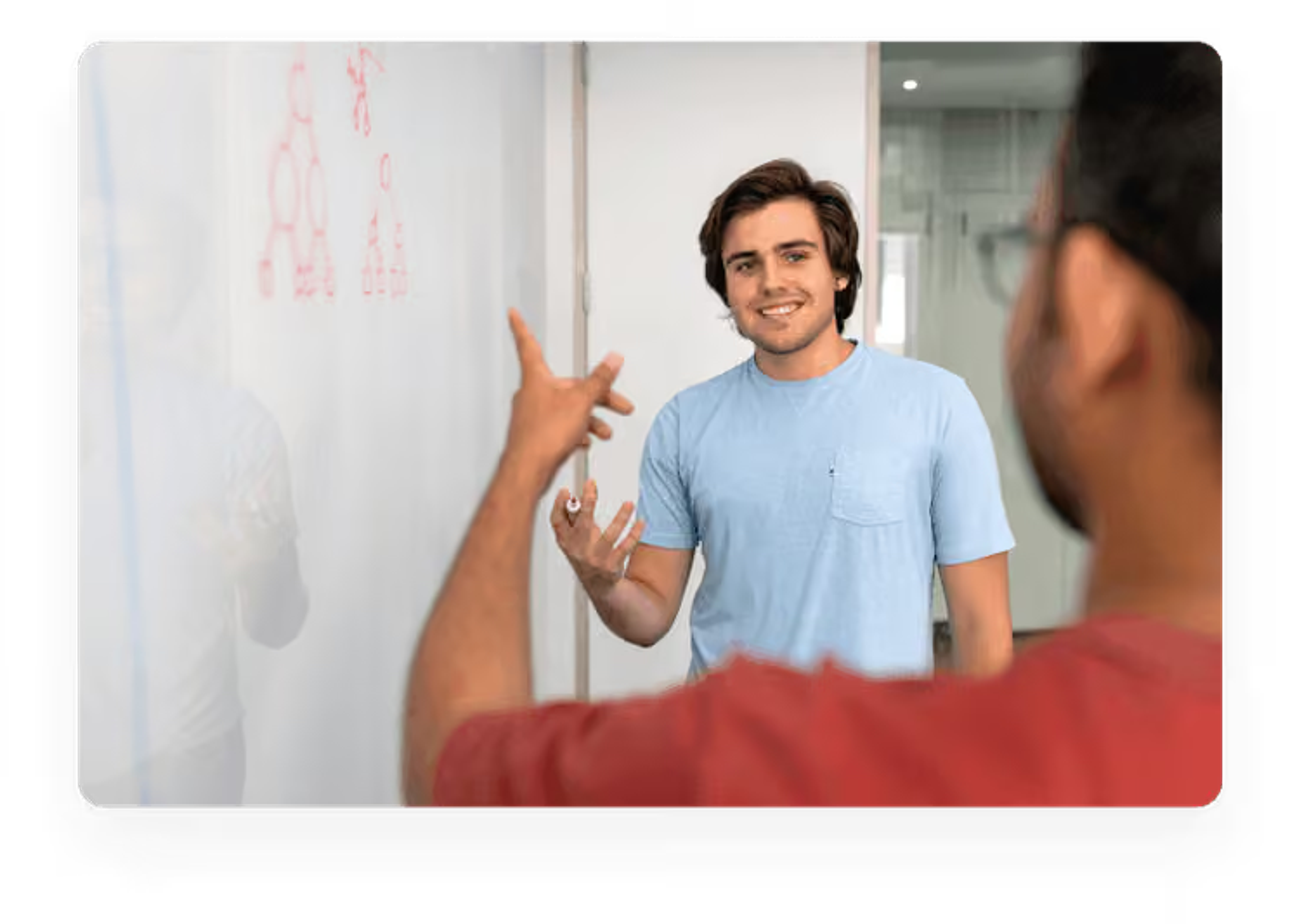 A young man in a blue t-shirt gesturing while explaining a concept by a whiteboard to a colleague