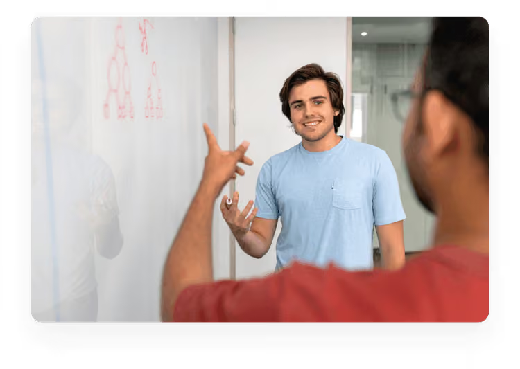 A young man in a blue t-shirt gesturing while explaining a concept by a whiteboard to a colleague