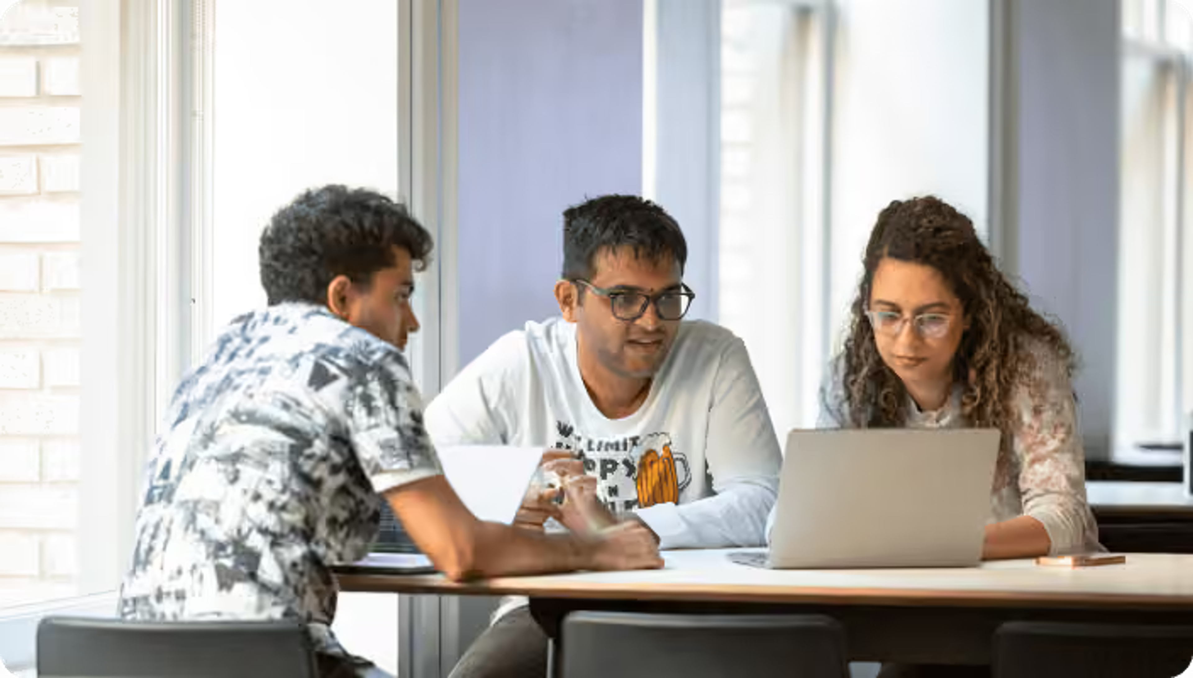 Three young professionals collaborating around a laptop at a table in a bright workspace.