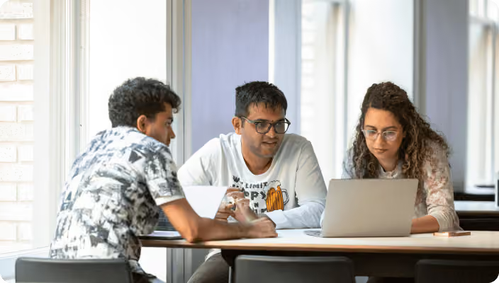 Three young professionals collaborating around a laptop at a table in a bright workspace.