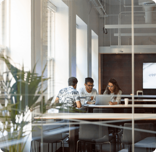Three colleagues collaborating at a table with a laptop in a bright, modern office viewed through a glass wall