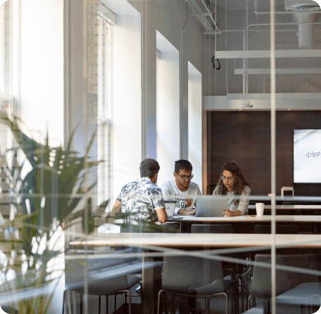 Three colleagues collaborating at a table with a laptop in a bright, modern office viewed through a glass wall