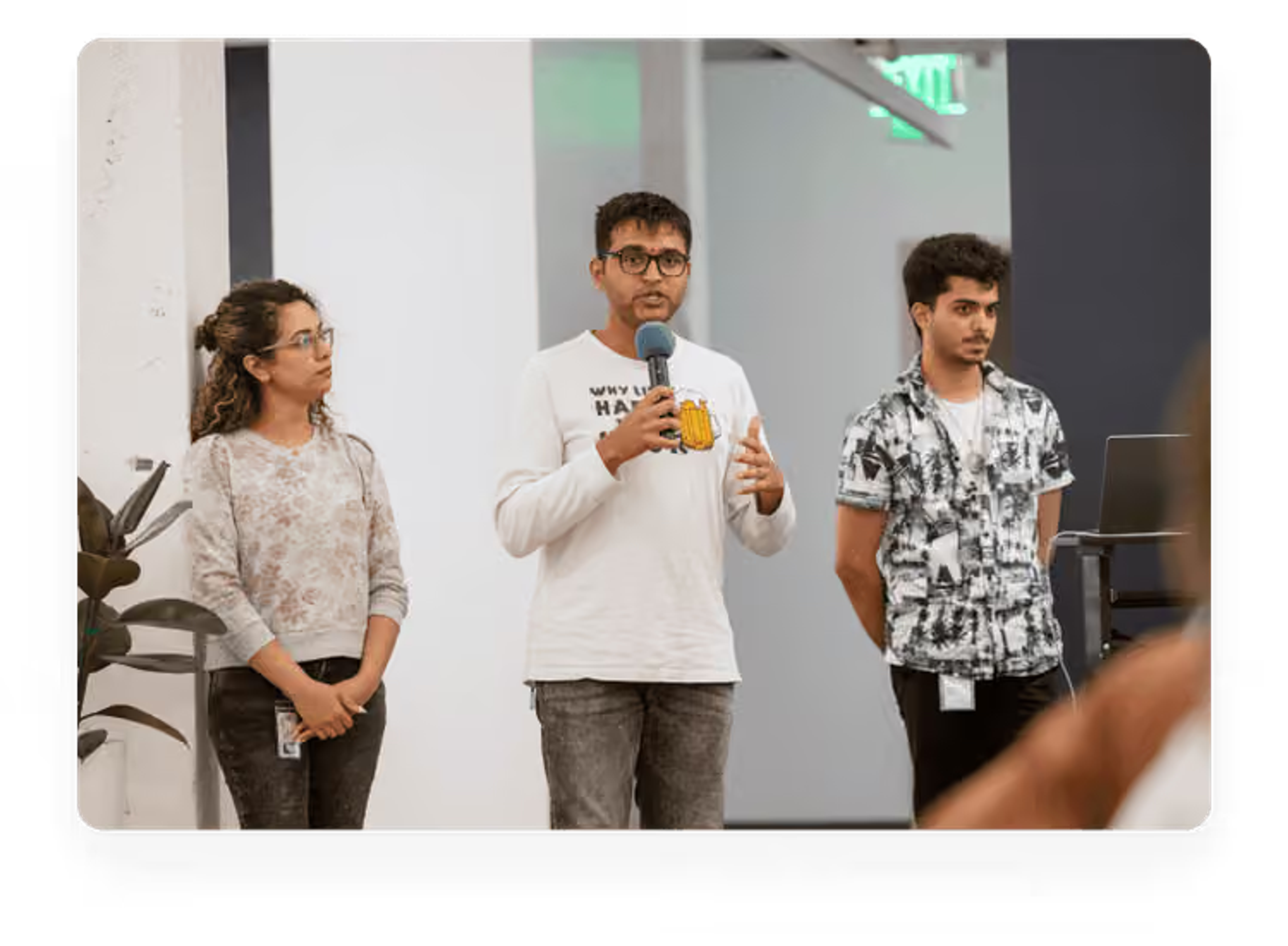 Three young colleagues standing together in a modern office space, with the person in the center speaking into a microphone