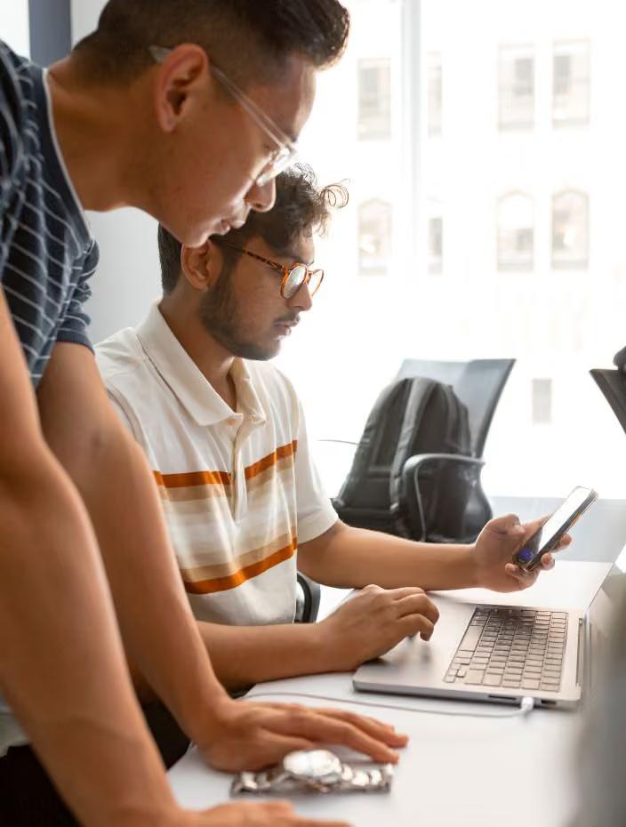 Two young men collaborating at a laptop in a bright, modern office setting, with one standing and one seated