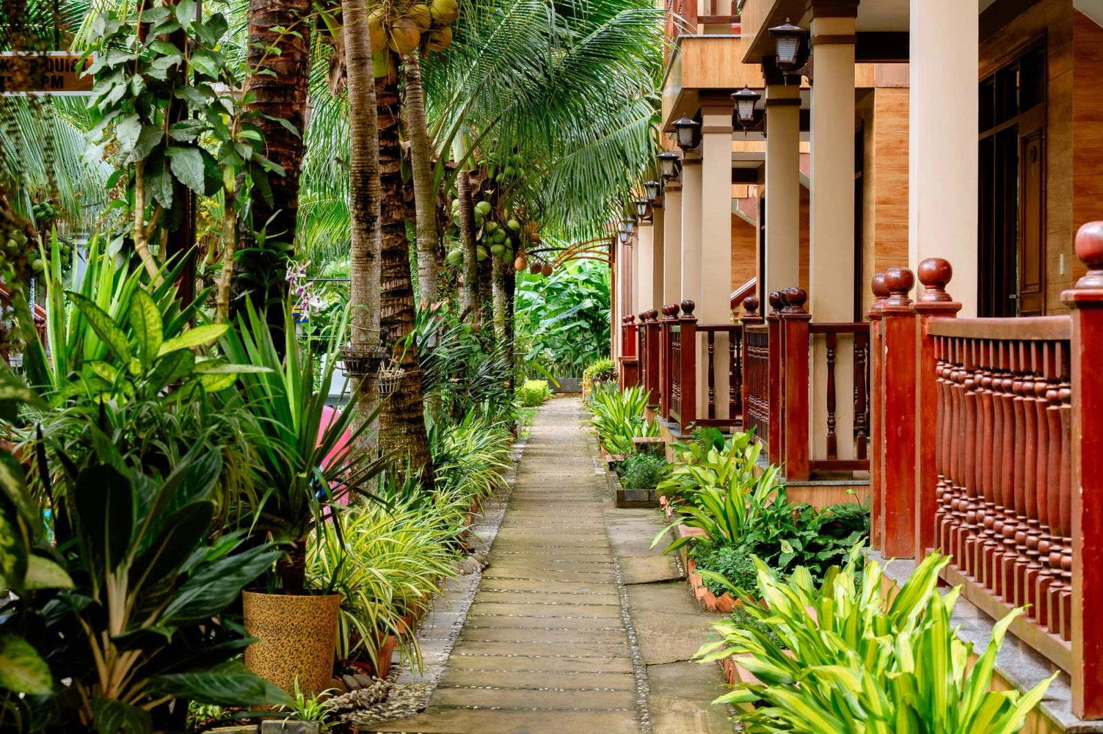 Garden walkway lined with tropical plants and covered verandas with red wooden railings.
