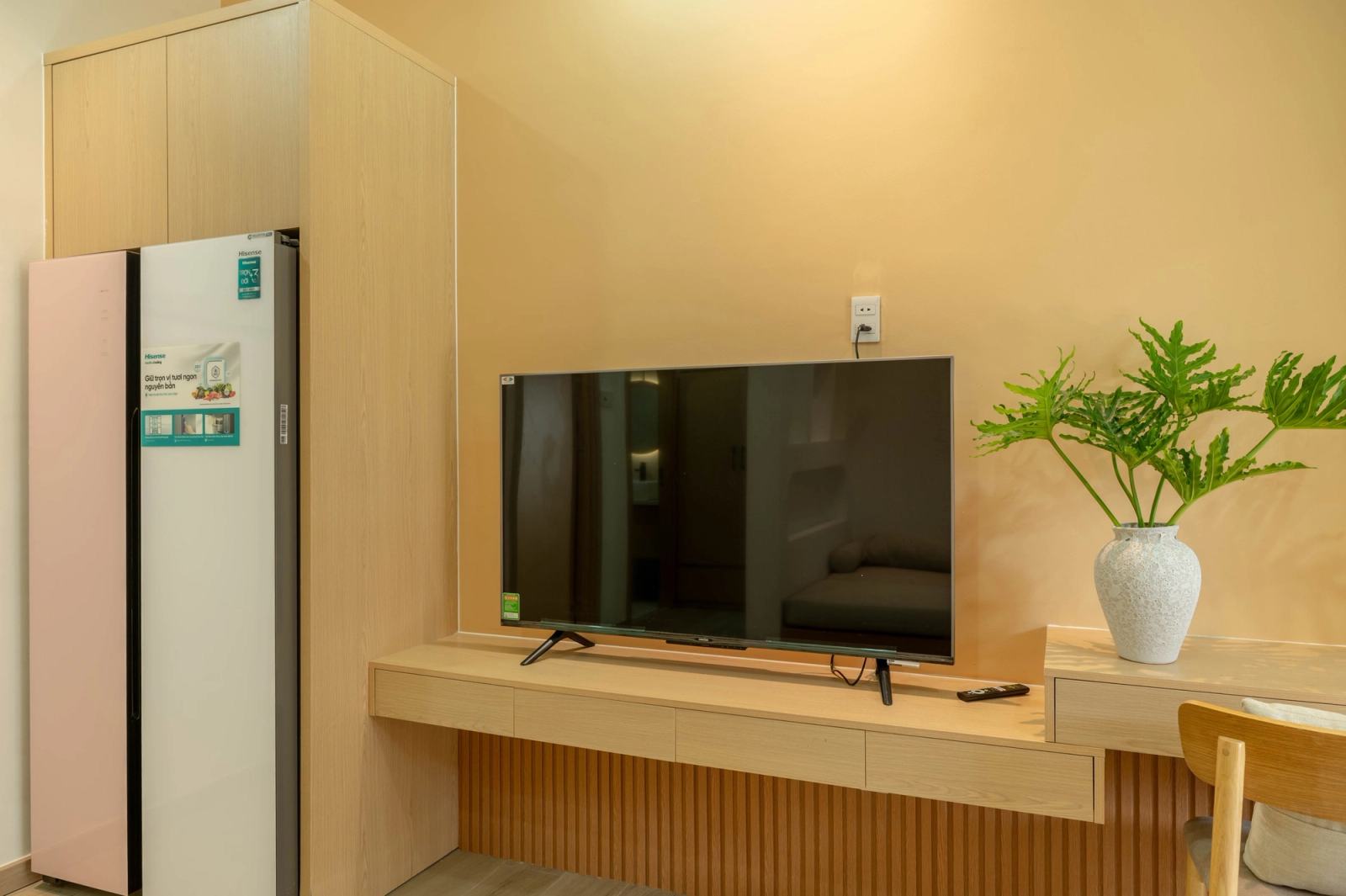 Television, refrigerator and a vase of greenery against a warm yellow wall.
