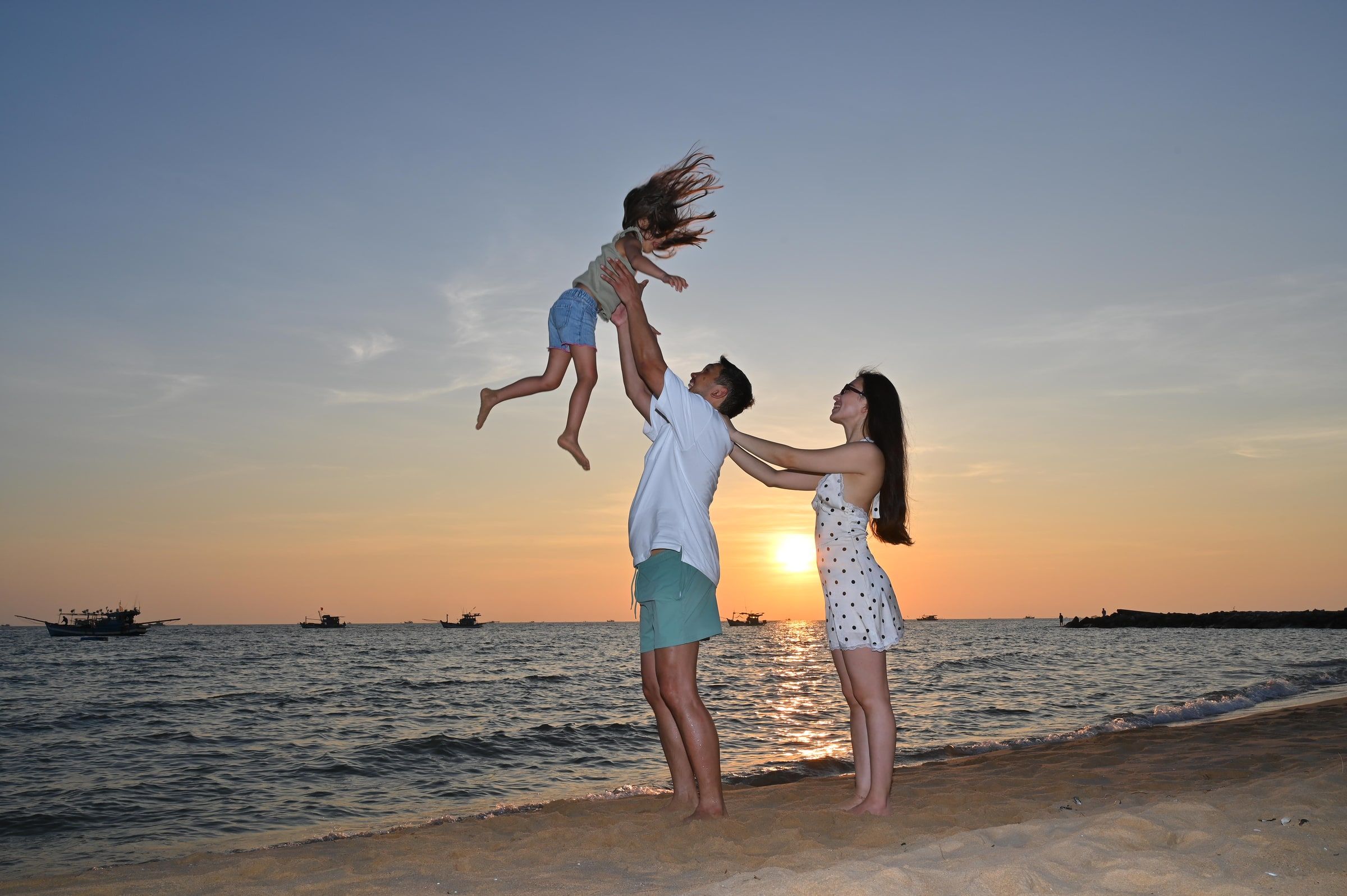 Parents playing with their child, lifted into the air on a beach at sunset.