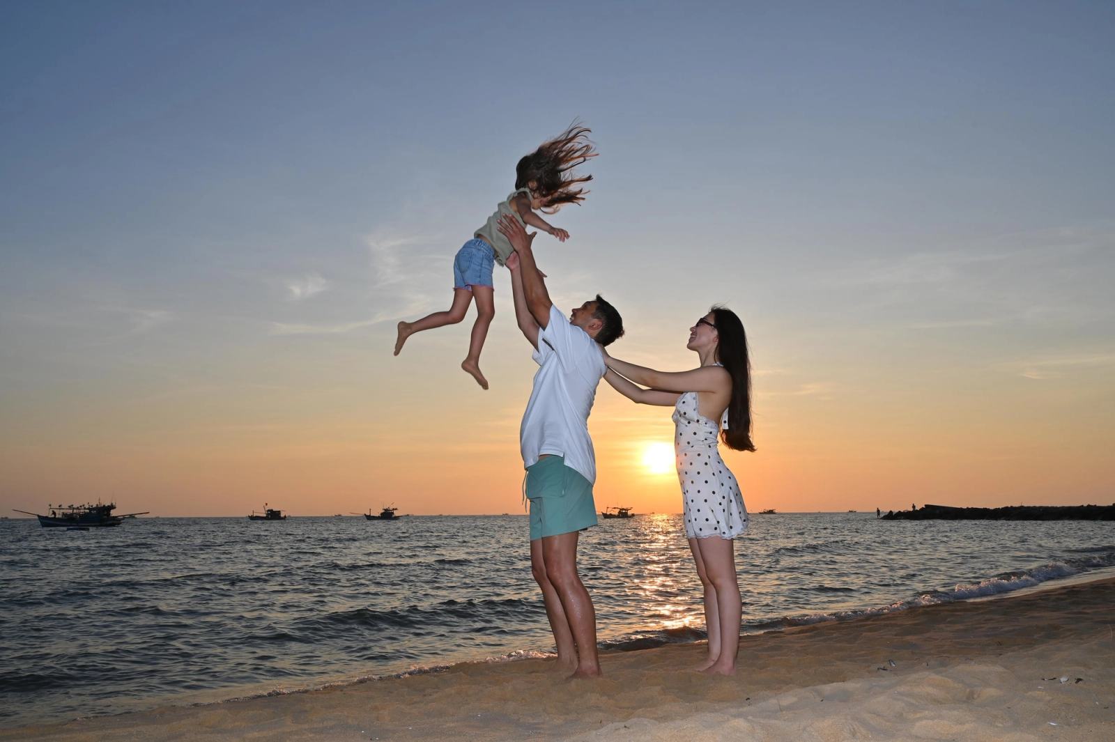Parents playing with their child, lifted into the air on a beach at sunset.