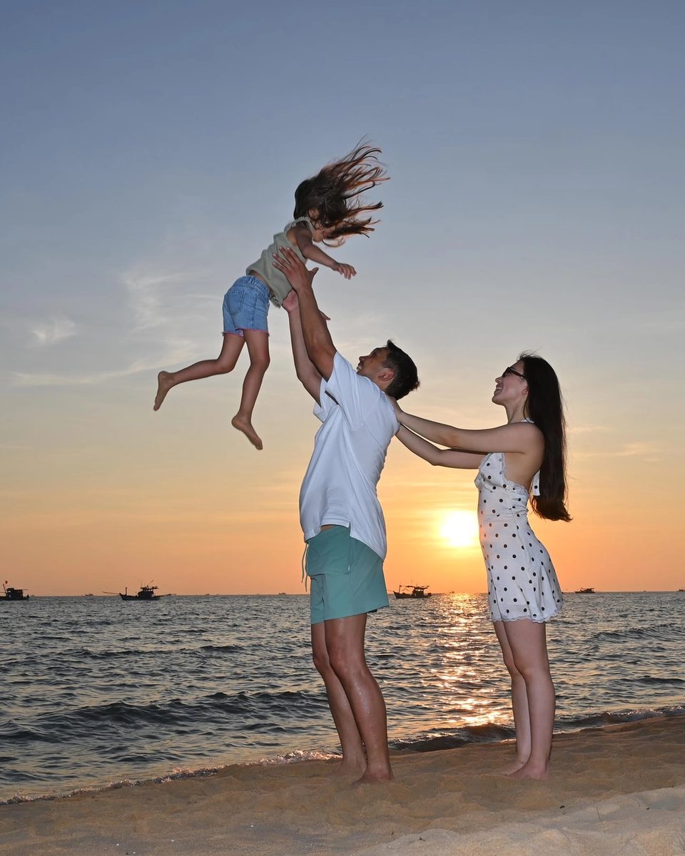 Parents playing with their child, lifted into the air on a beach at sunset.