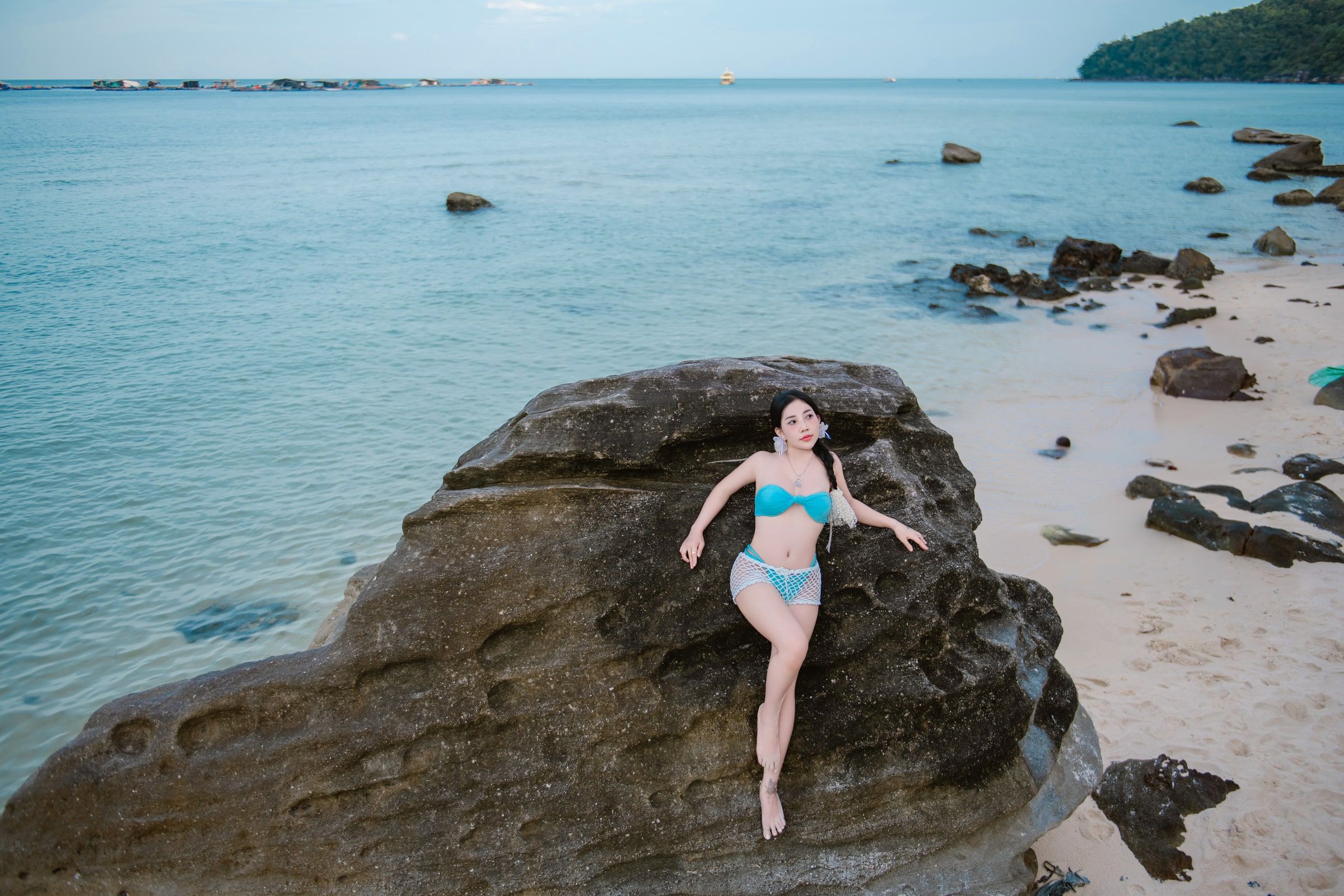 Woman in blue swimwear seated on coastal rocks beside a calm sea.