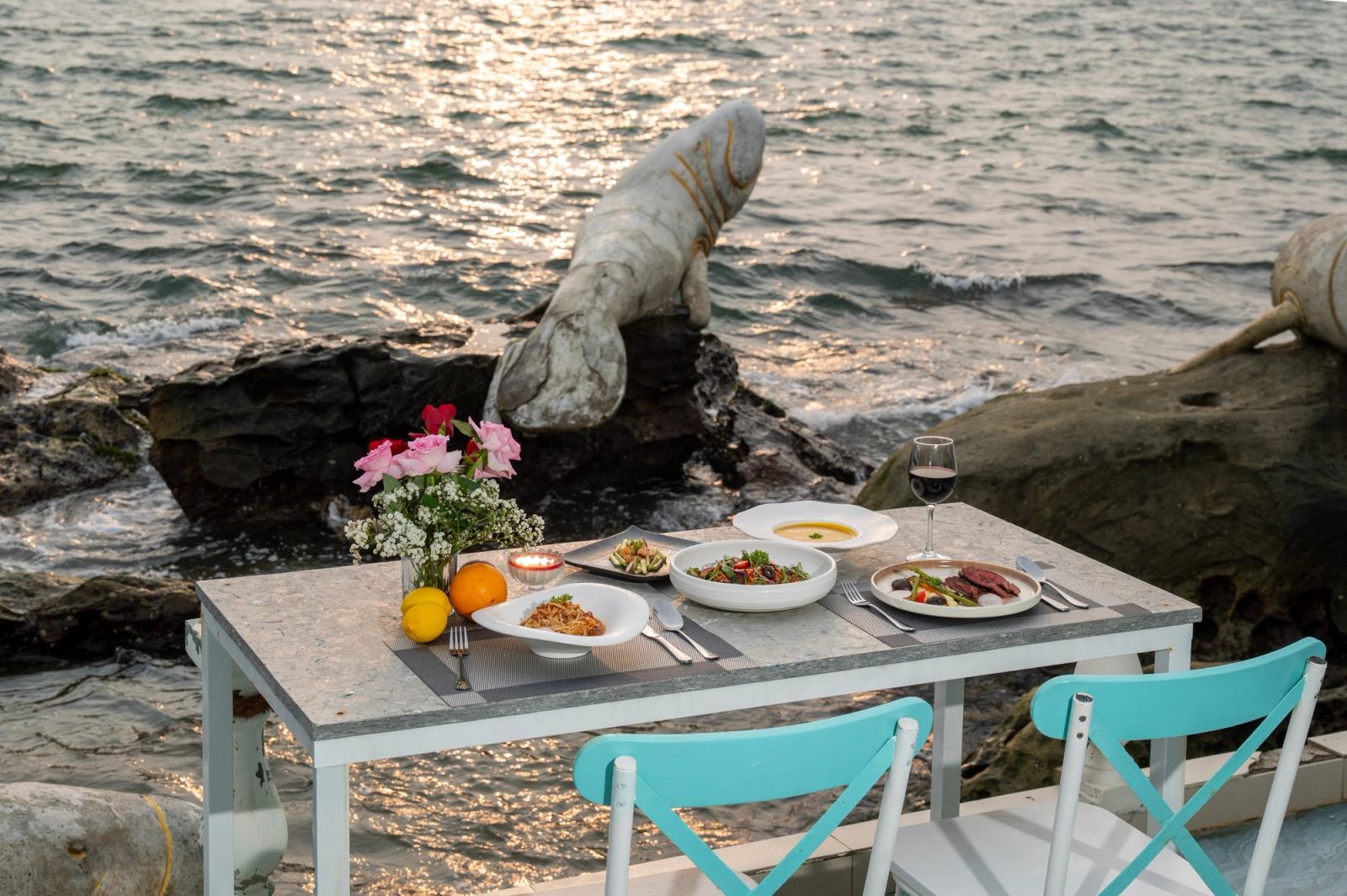 A table set with dishes and flowers beside turquoise chairs overlooking the rocky shoreline.