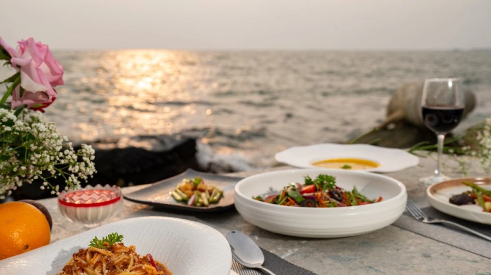Plated dishes and a glass of red wine on a seaside table in warm sunset light.