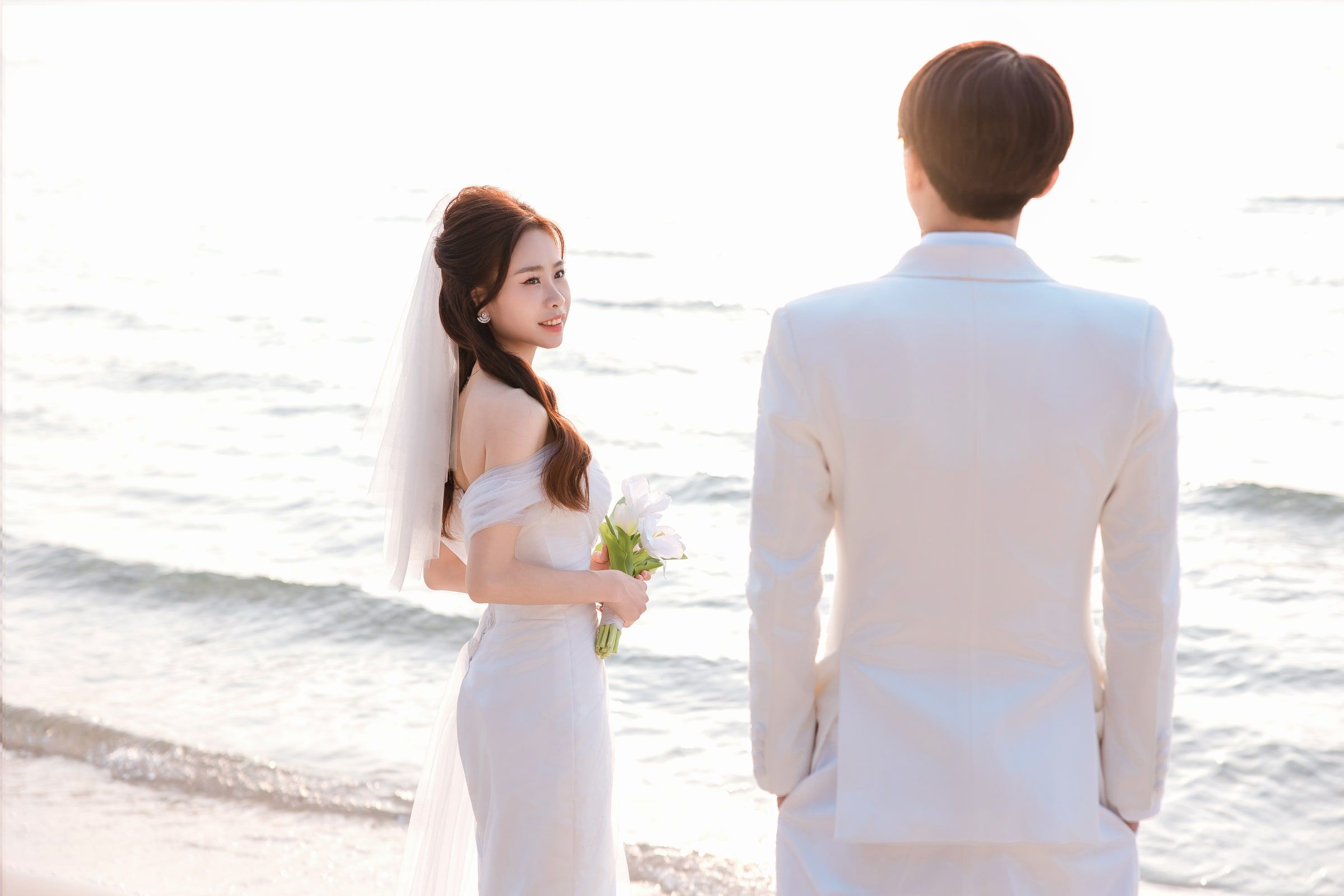 Bride holding a white bouquet and looking towards her partner beside the sea.