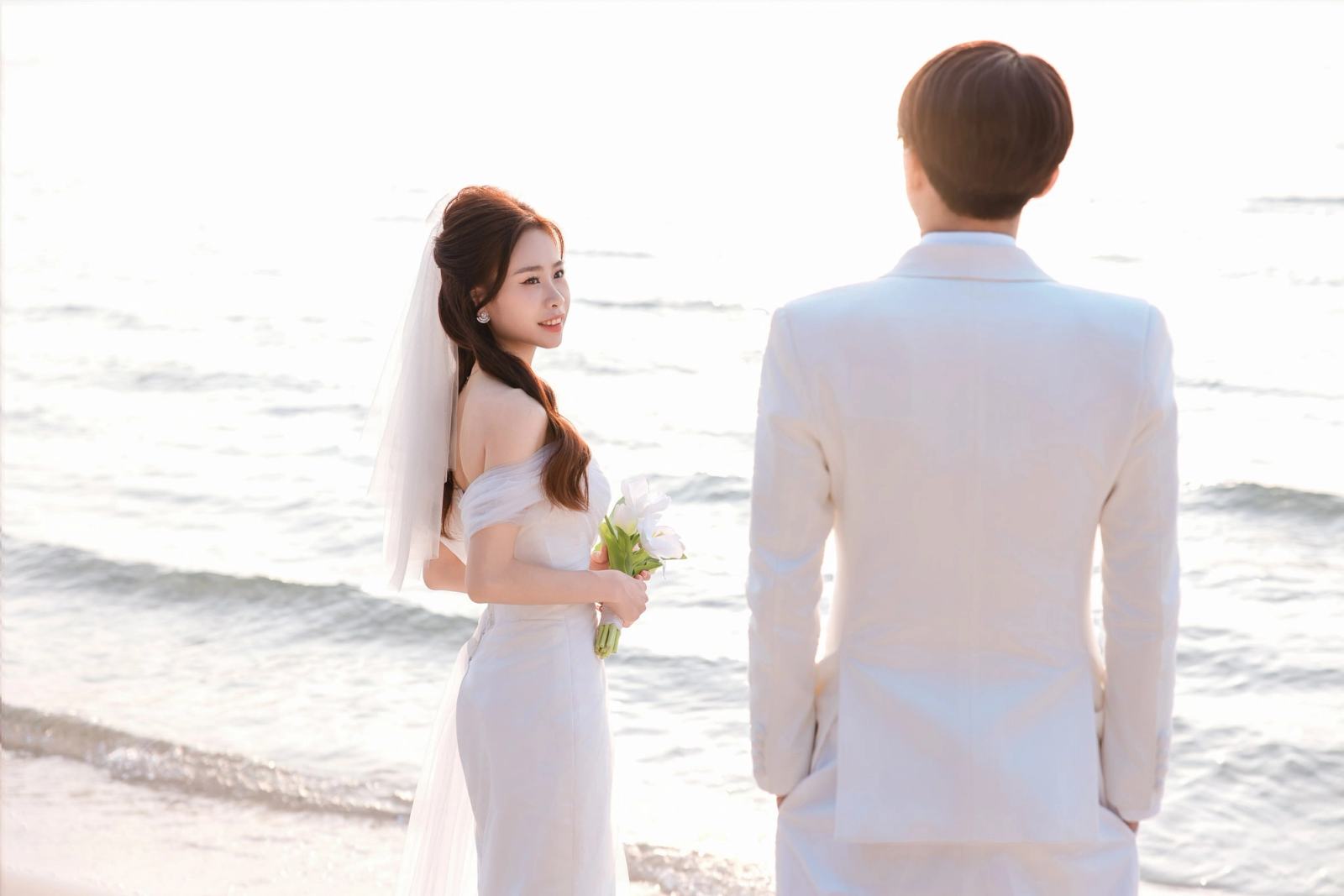 Bride holding a white bouquet and looking towards her partner beside the sea.