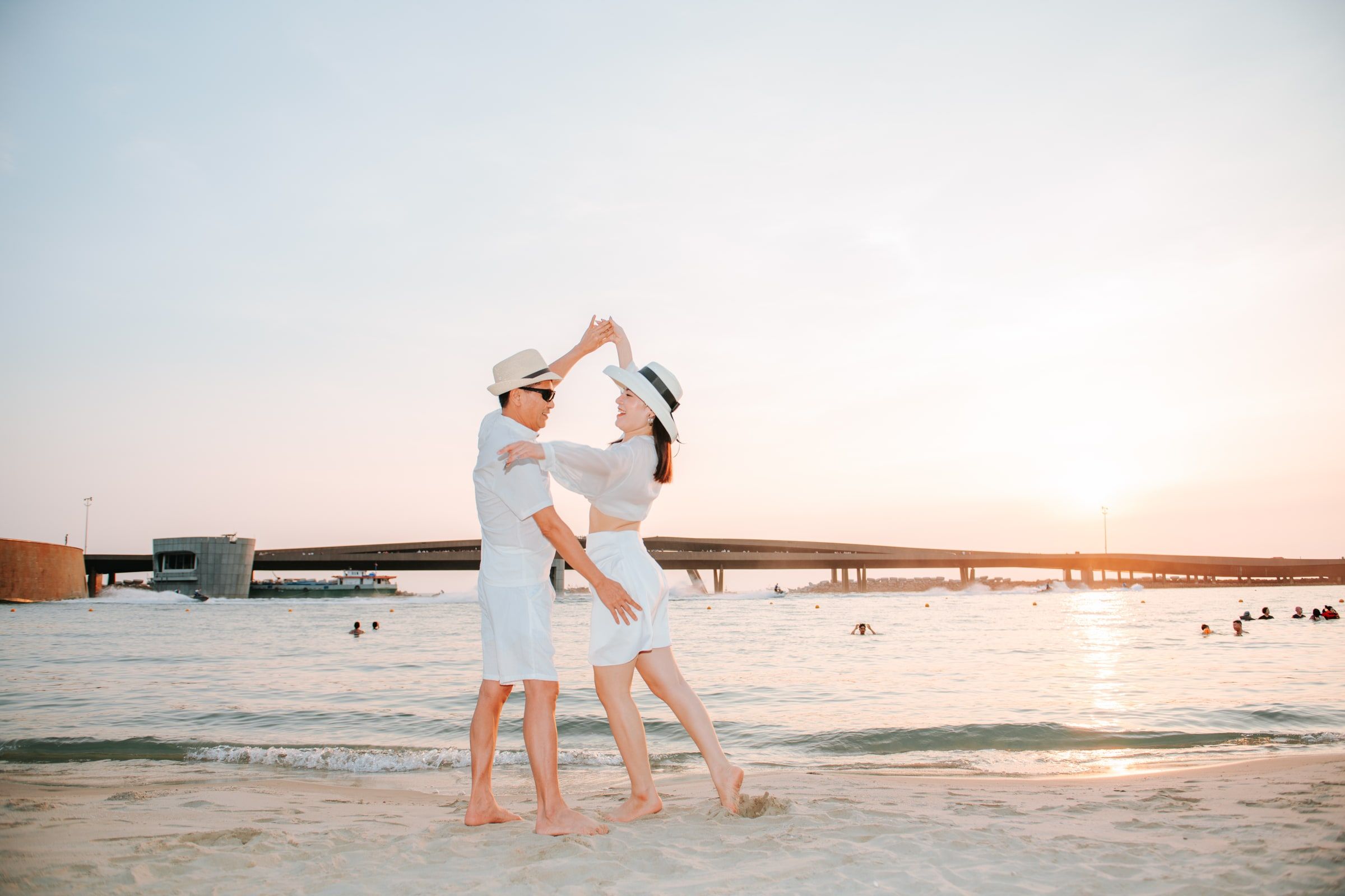 Couple in white outfits and sun hats dancing at the water's edge at sunset.