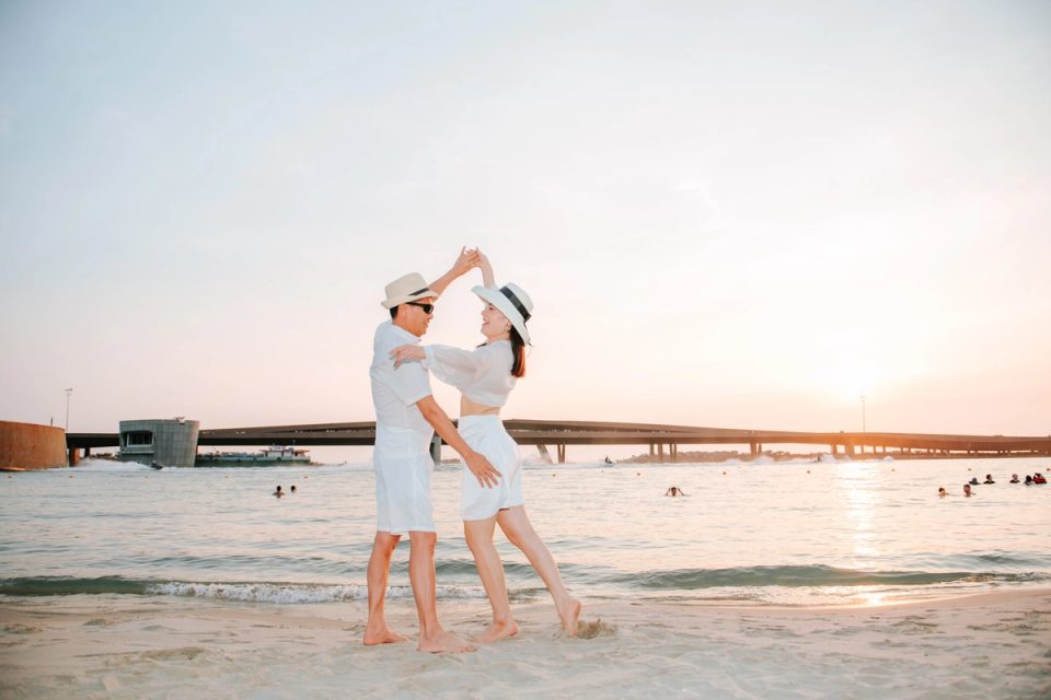 Couple in white outfits and sun hats dancing at the water's edge at sunset.
