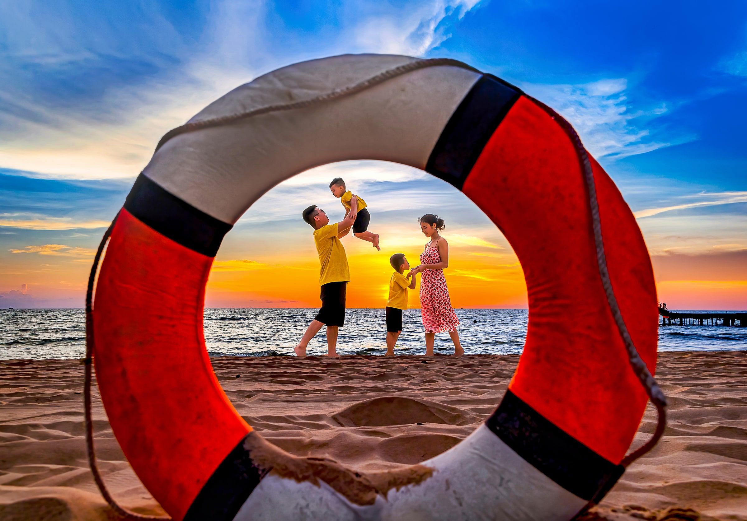 Family playing on the beach at sunset, framed through a red-and-white lifebuoy.