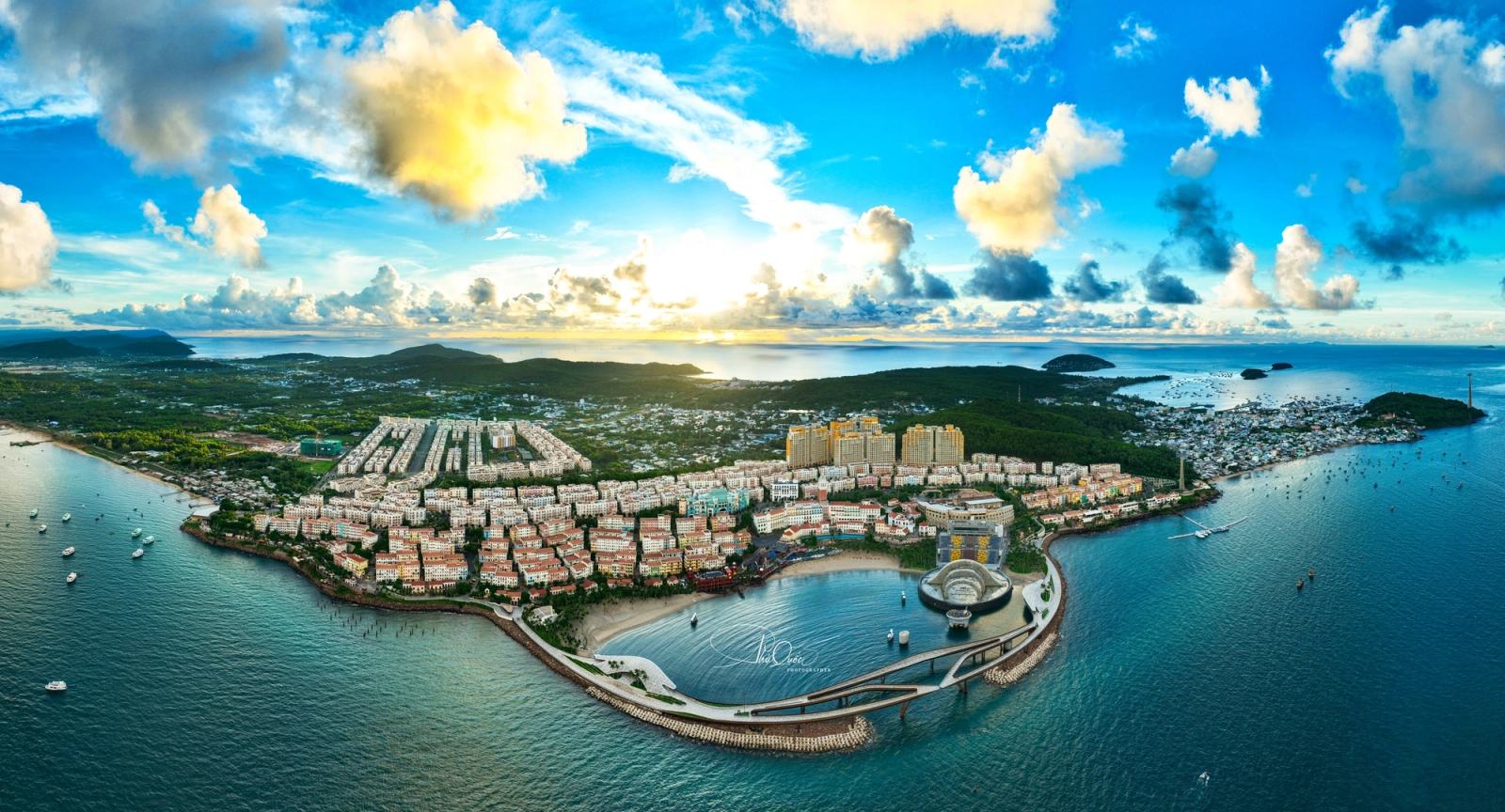 Panoramic aerial view of Sunset Town, Kiss Bridge and the curved waterfront beneath sunlit clouds.