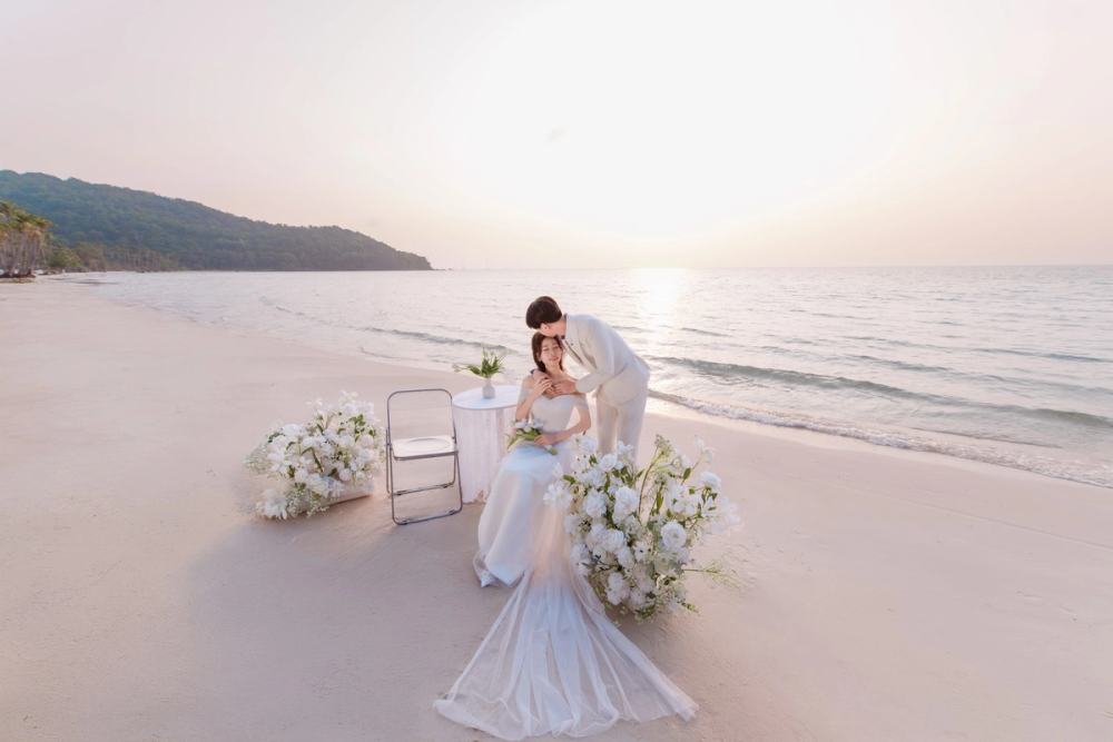 Couple in wedding attire beside a white floral arrangement on a quiet beach in soft evening light.