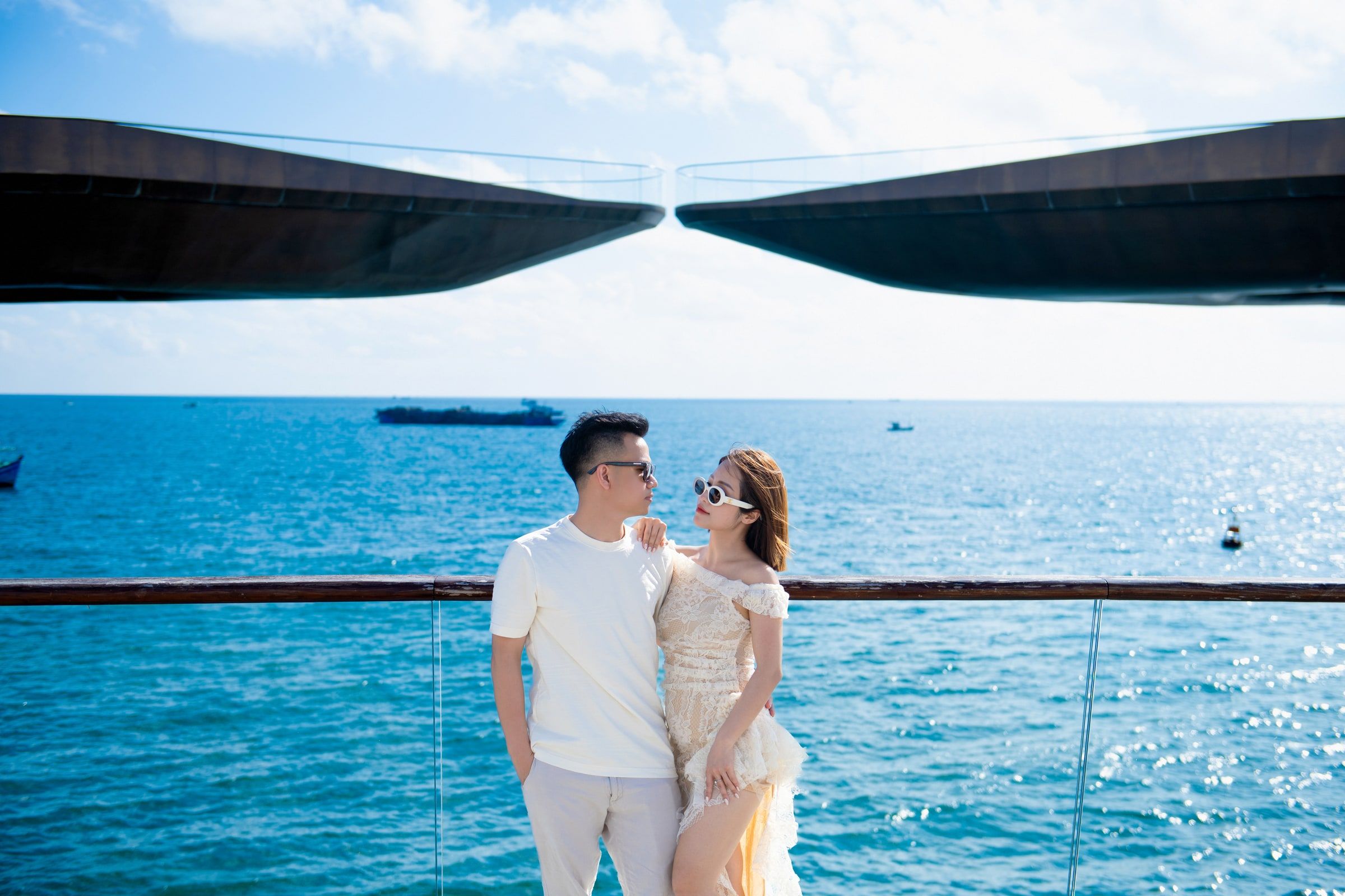 Couple standing together beside a waterfront railing with the two spans of Kiss Bridge behind them.