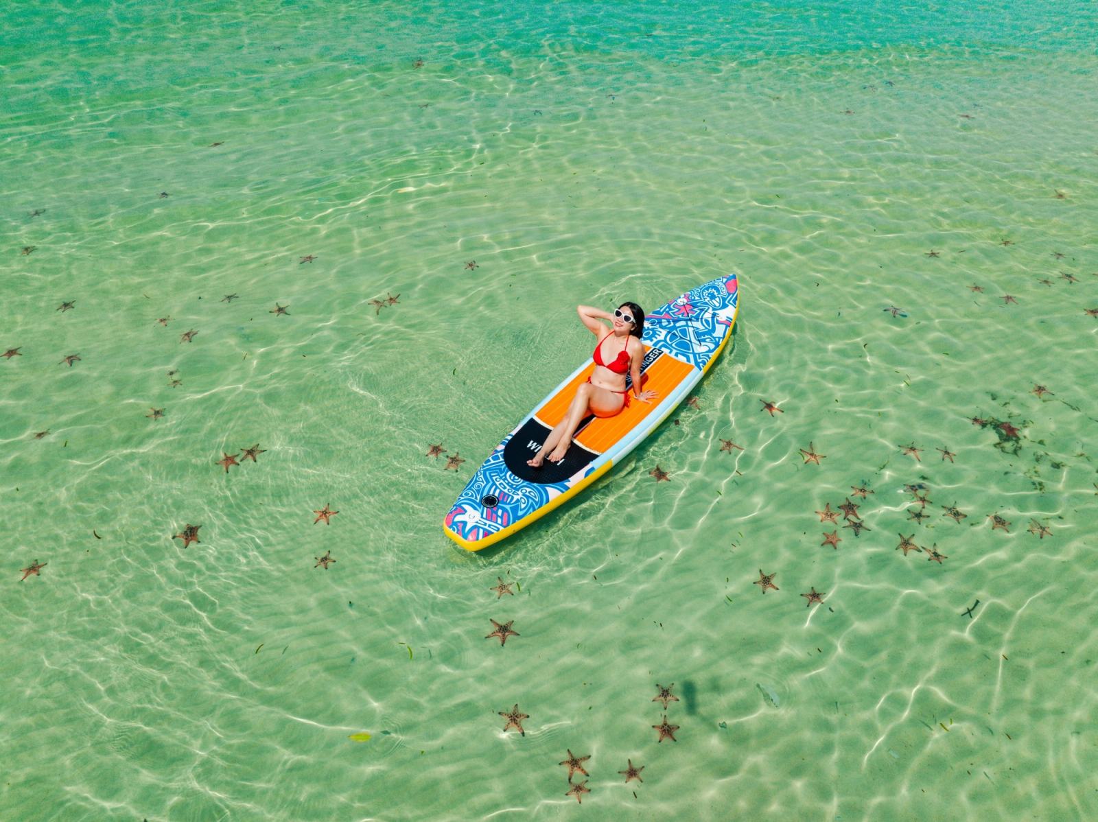 Overhead portrait of a woman seated on a paddleboard in clear shallow water.