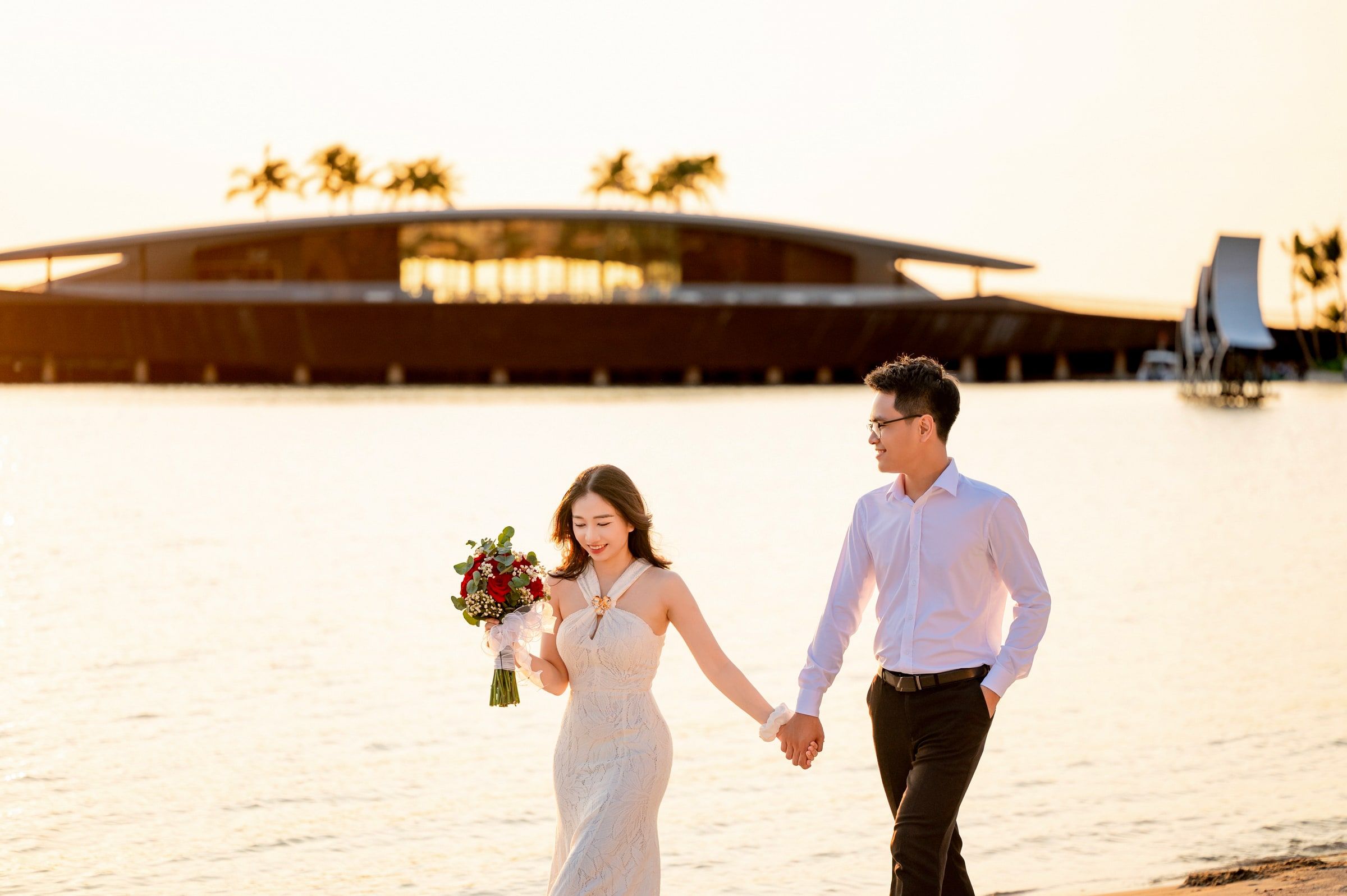 Couple holding hands beside the sea in warm evening light, the woman carrying flowers.