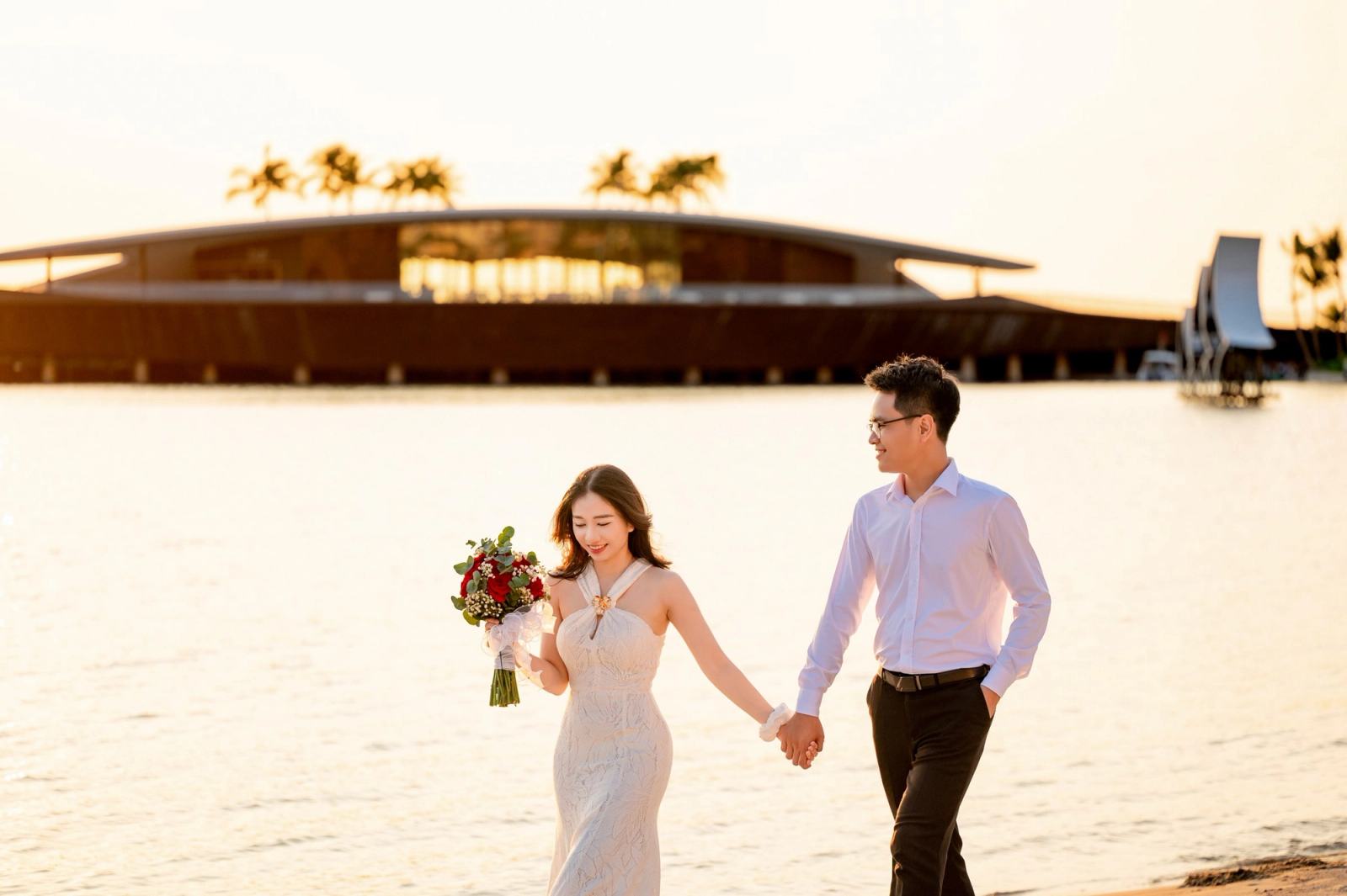 Couple holding hands beside the sea in warm evening light, the woman carrying flowers.