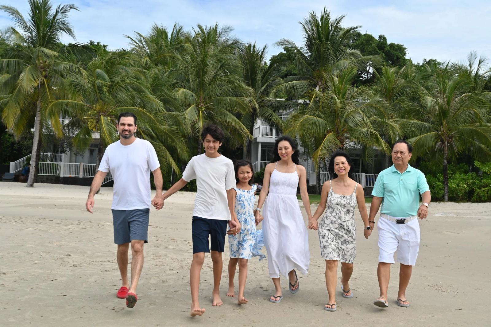 Family members walking hand in hand along a beach in front of palm trees.