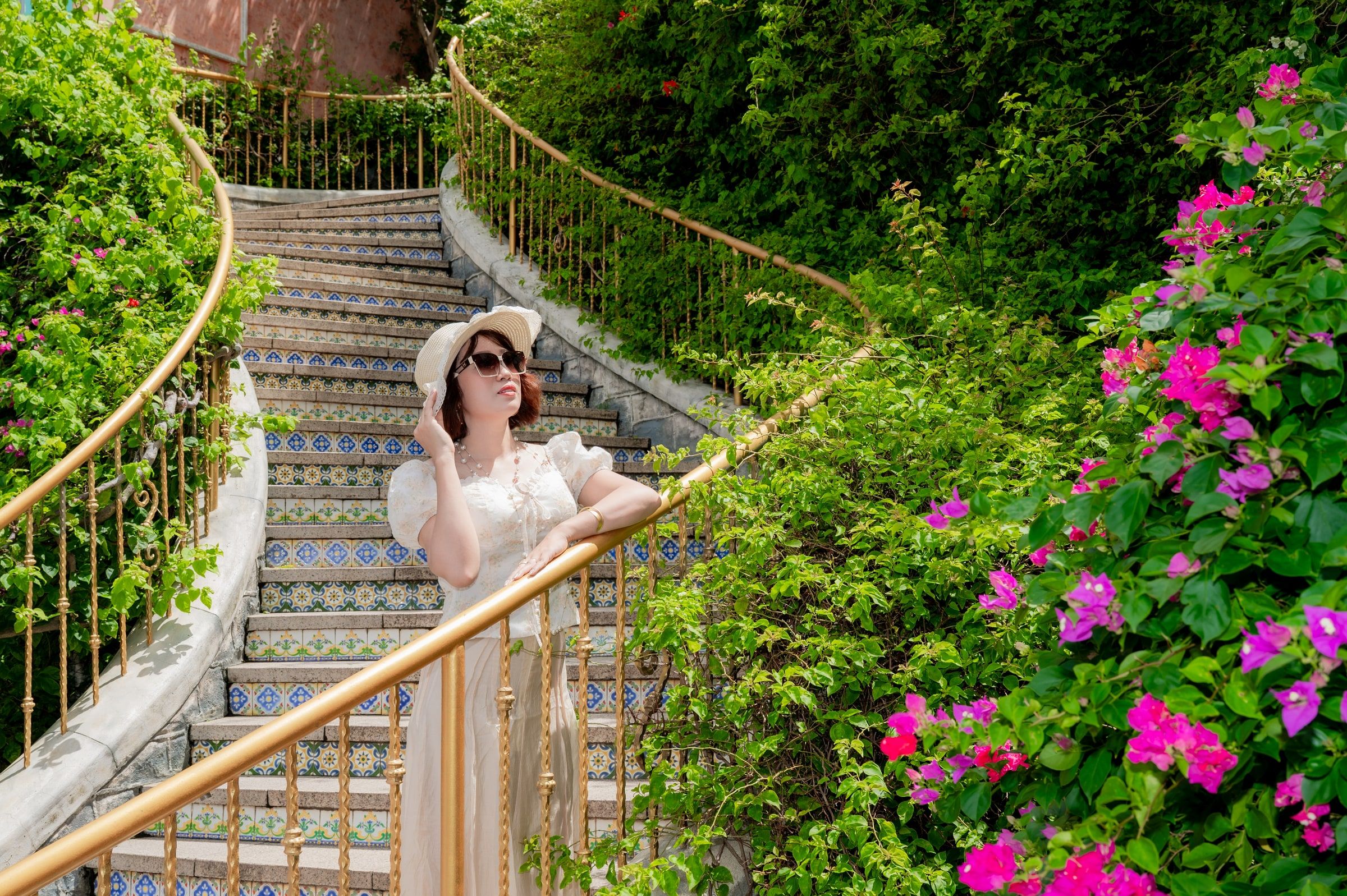 Woman in a sun hat beside a garden staircase framed by green foliage and pink flowers.