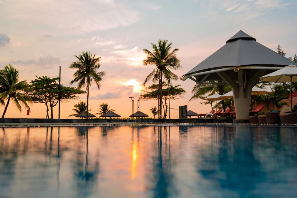 Sunset reflected in a resort pool beside palm trees and a shaded pavilion.