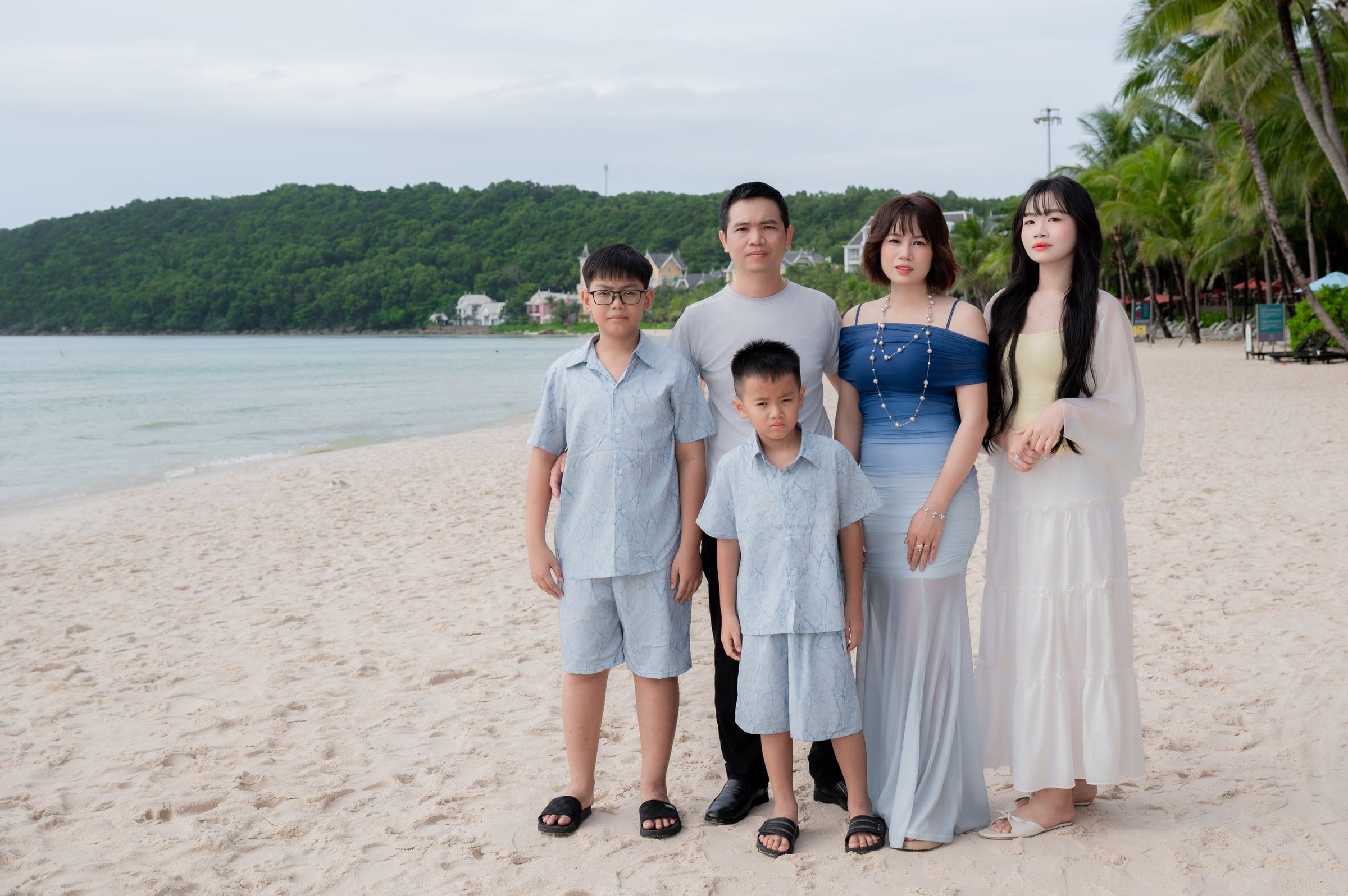 Family of five posing on a beach with palm trees and green hills behind them.