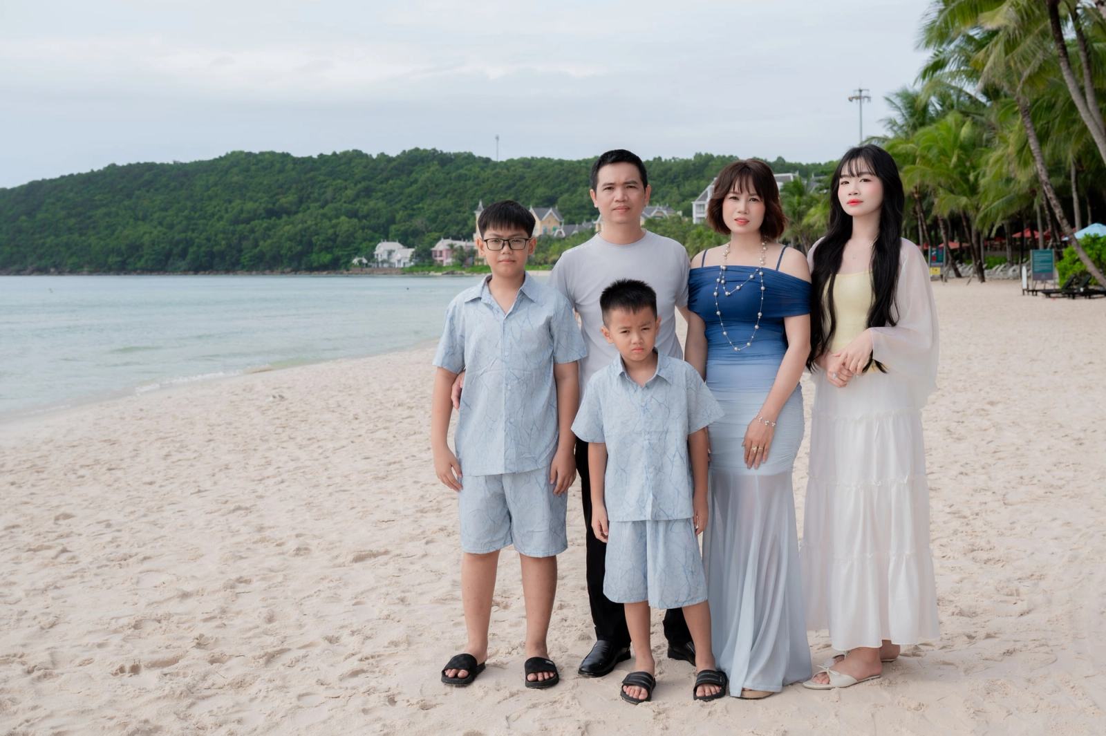 Family of five posing on a beach with palm trees and green hills behind them.