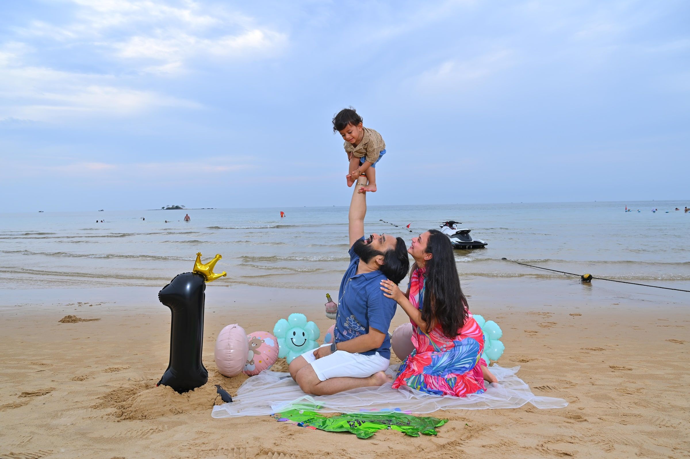 Parents playing with a young child at a beach picnic with a number-one balloon.
