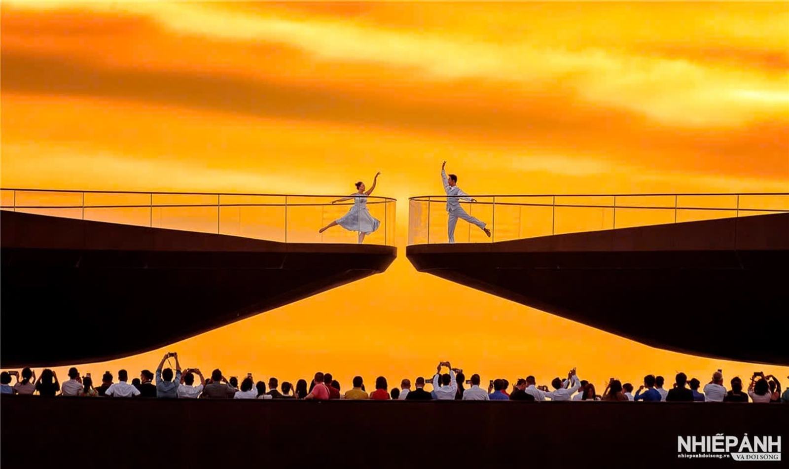 Two performers on opposite sides of Kiss Bridge against an orange sky, with spectators below.
