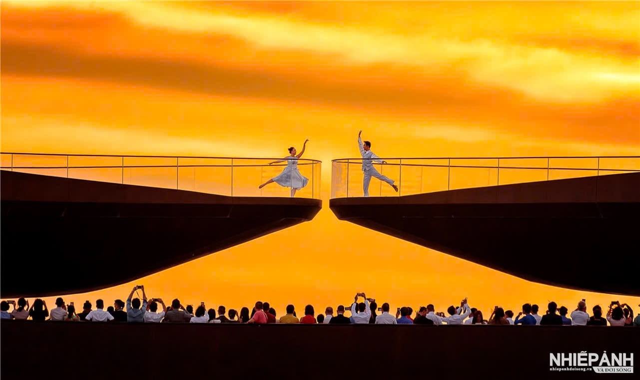 Two performers on opposite sides of Kiss Bridge against an orange sky, with spectators below.