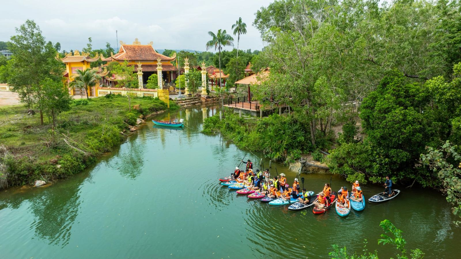 A group of kayaks on a calm river beside trees and an ornate riverside building.