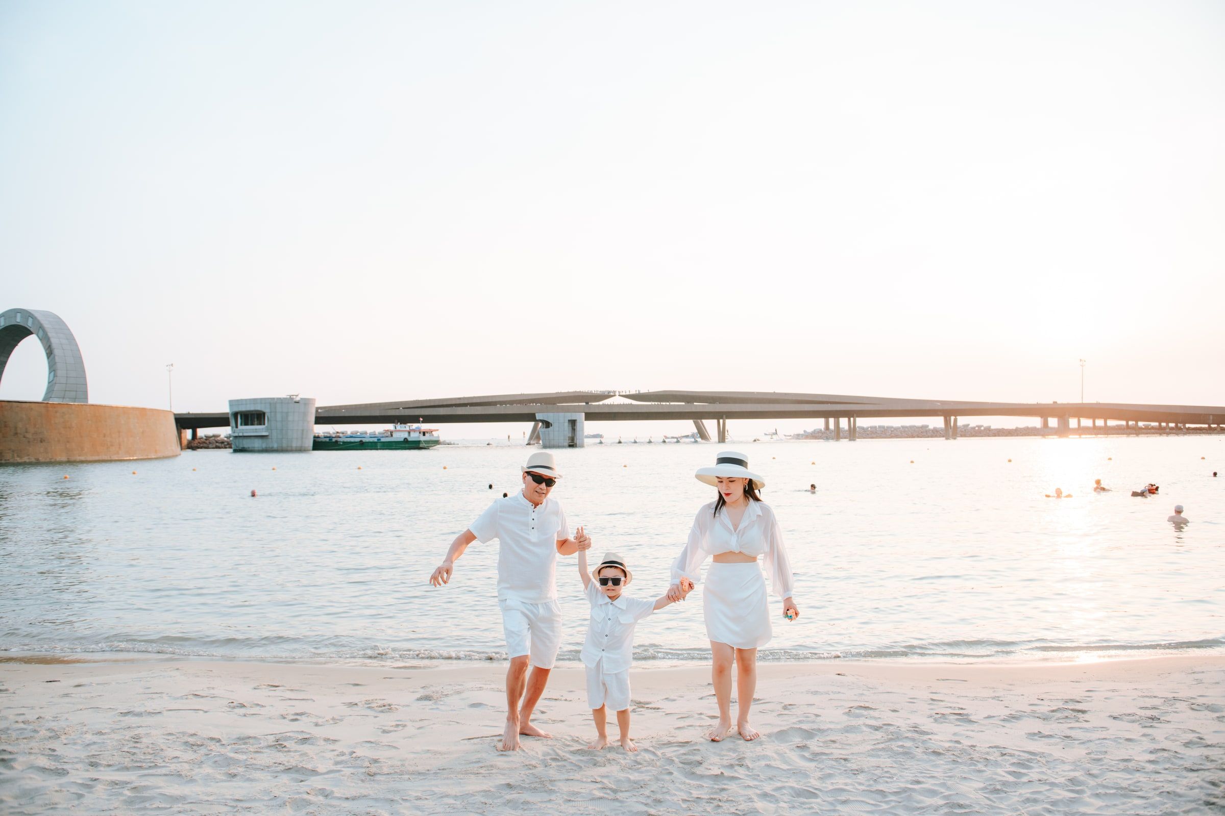 Parents holding their child's hands as they walk along the shoreline in white outfits.
