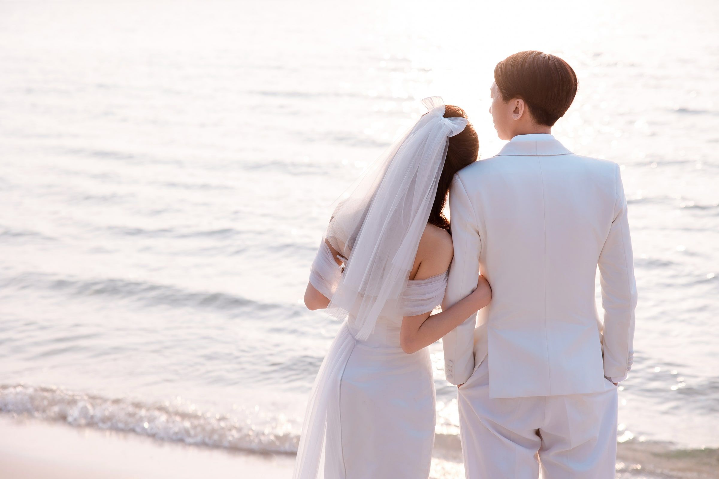 Bride resting her head on her partner's shoulder as they face the sea, seen from behind.