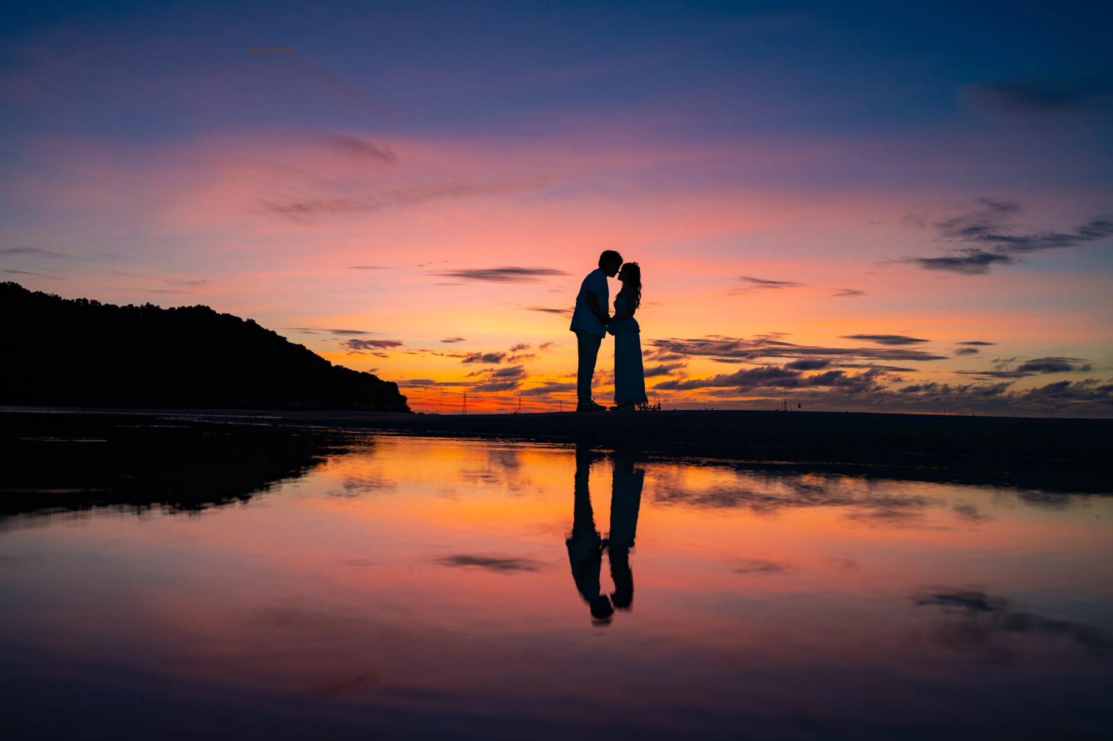 Silhouette of a couple standing close together at sunset, reflected in shallow water.