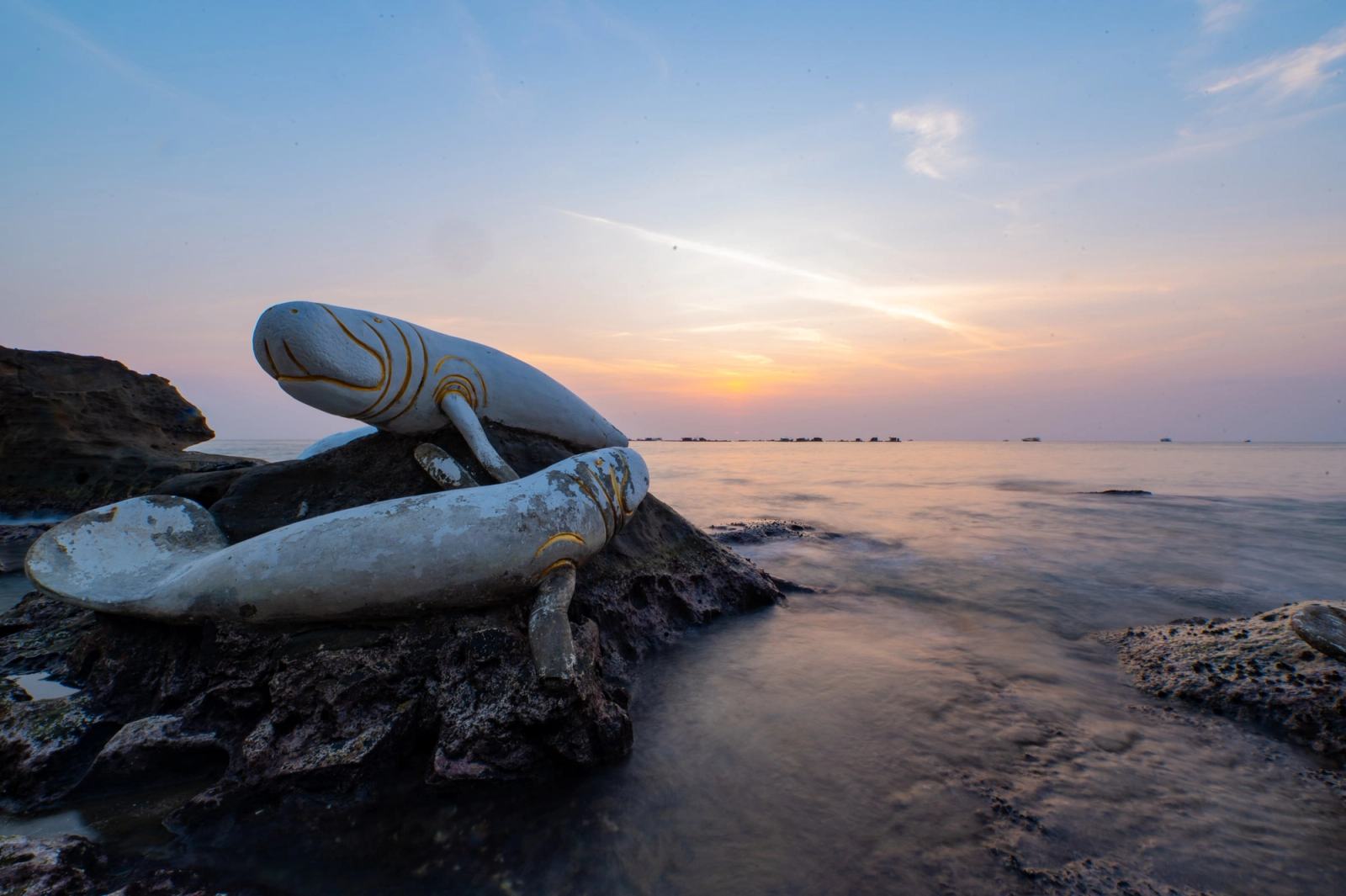 Fish sculptures on coastal rocks beneath a pastel evening sky.