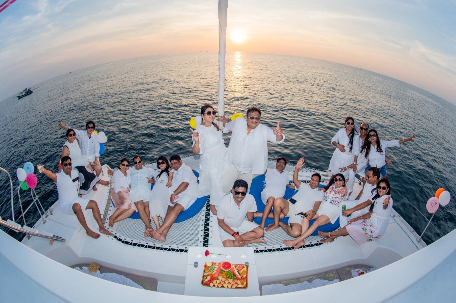 Guests gathered on a catamaran deck at sunset with balloons and a fruit platter.