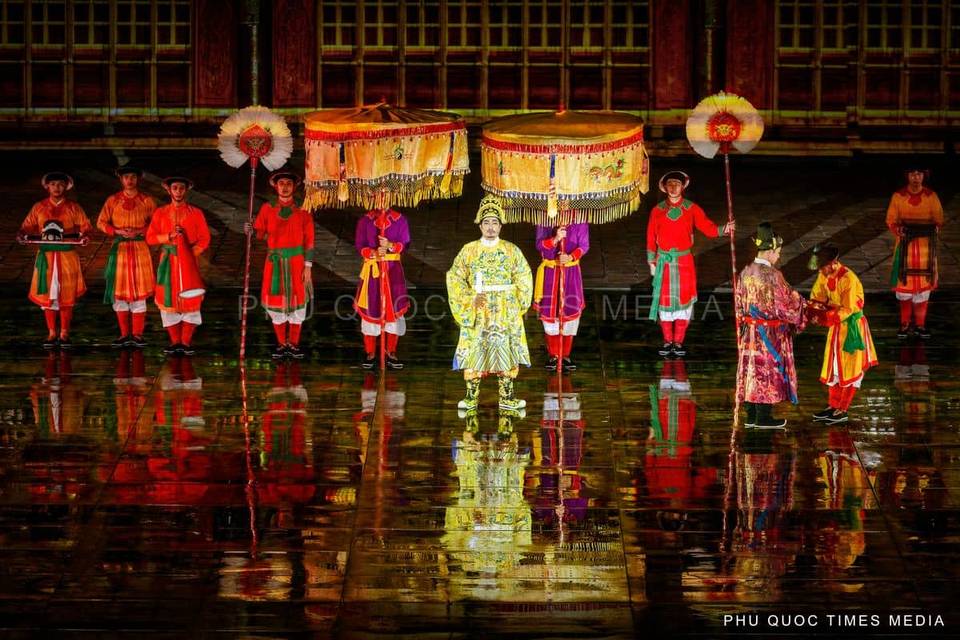 Performers in colourful ceremonial costumes beneath decorative canopies, reflected on a glossy stage