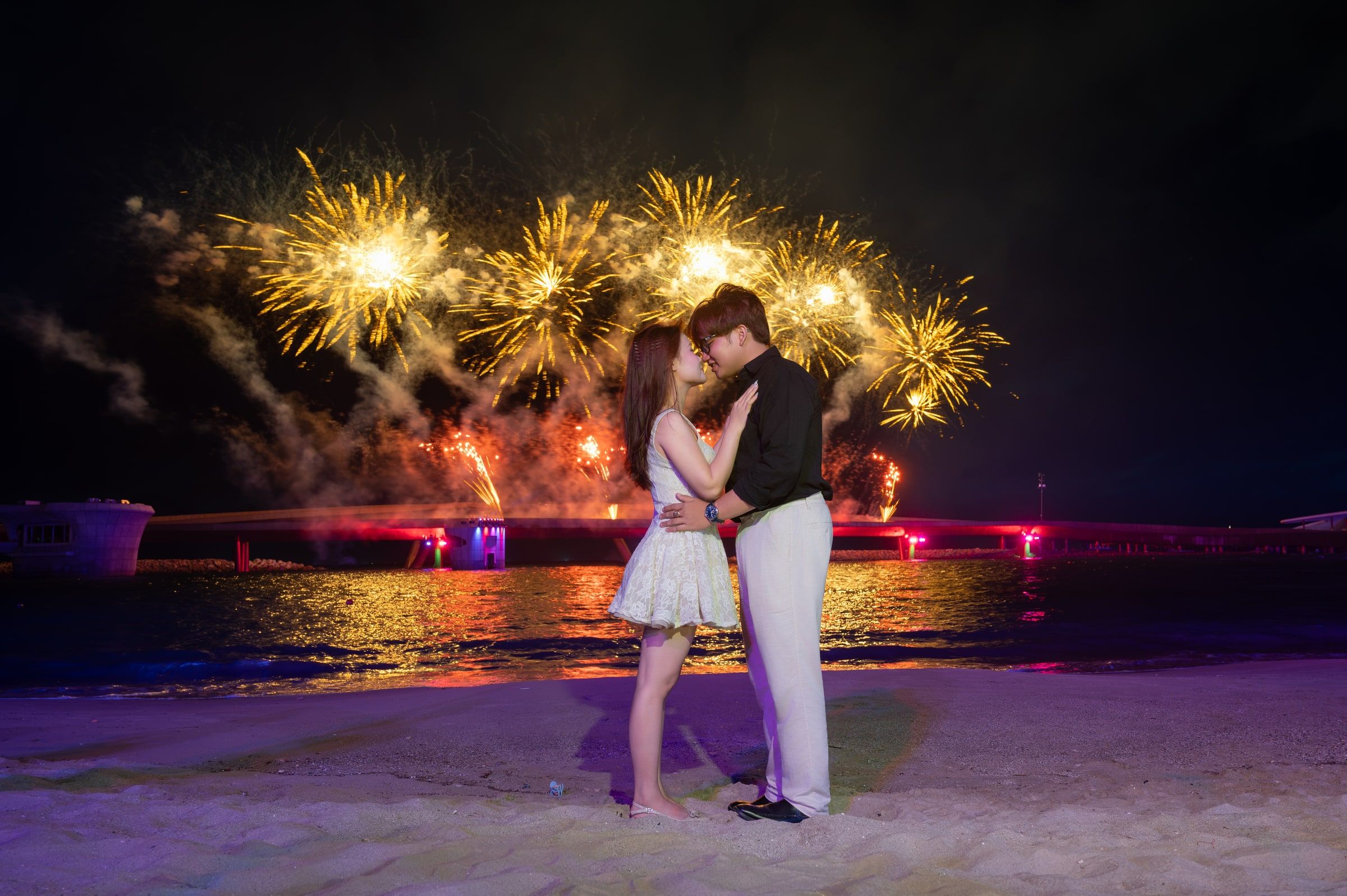 Couple kissing beside the waterfront beneath golden fireworks at night.