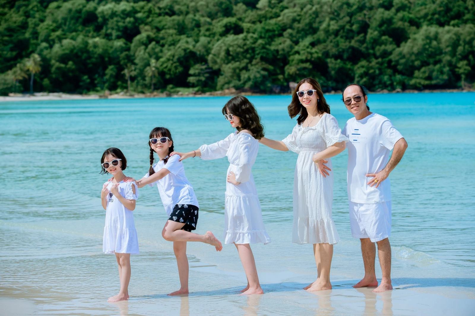 Family playing together in shallow turquoise water beside a tree-lined shore.
