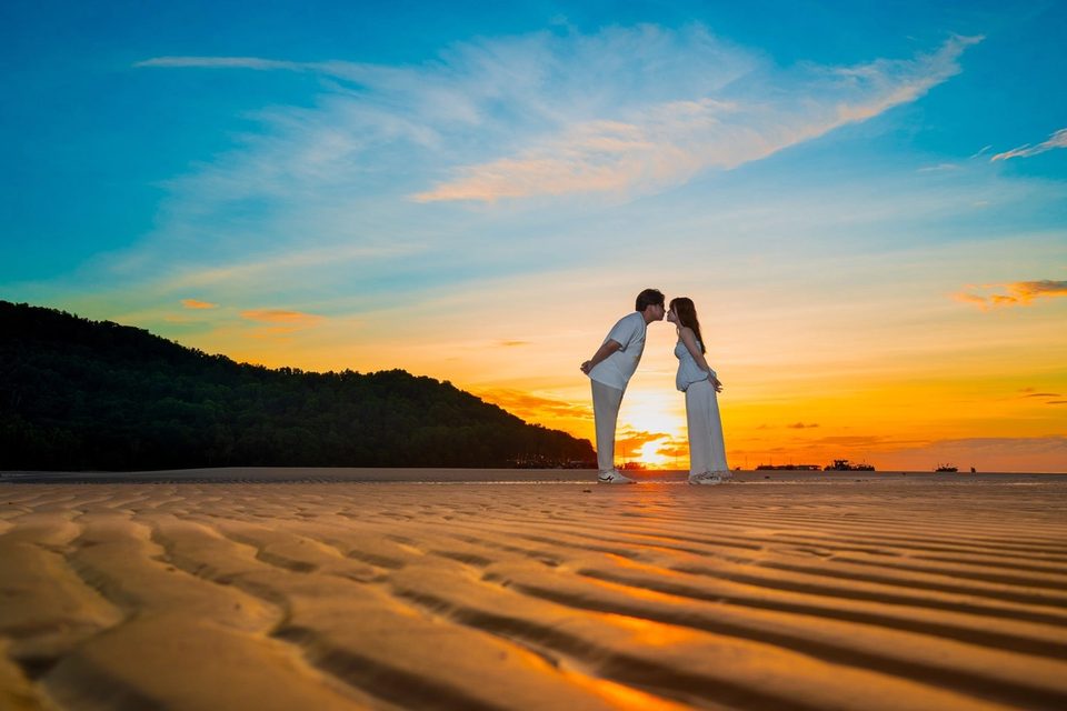 A couple kissing on rippled sand beneath an evening sky.