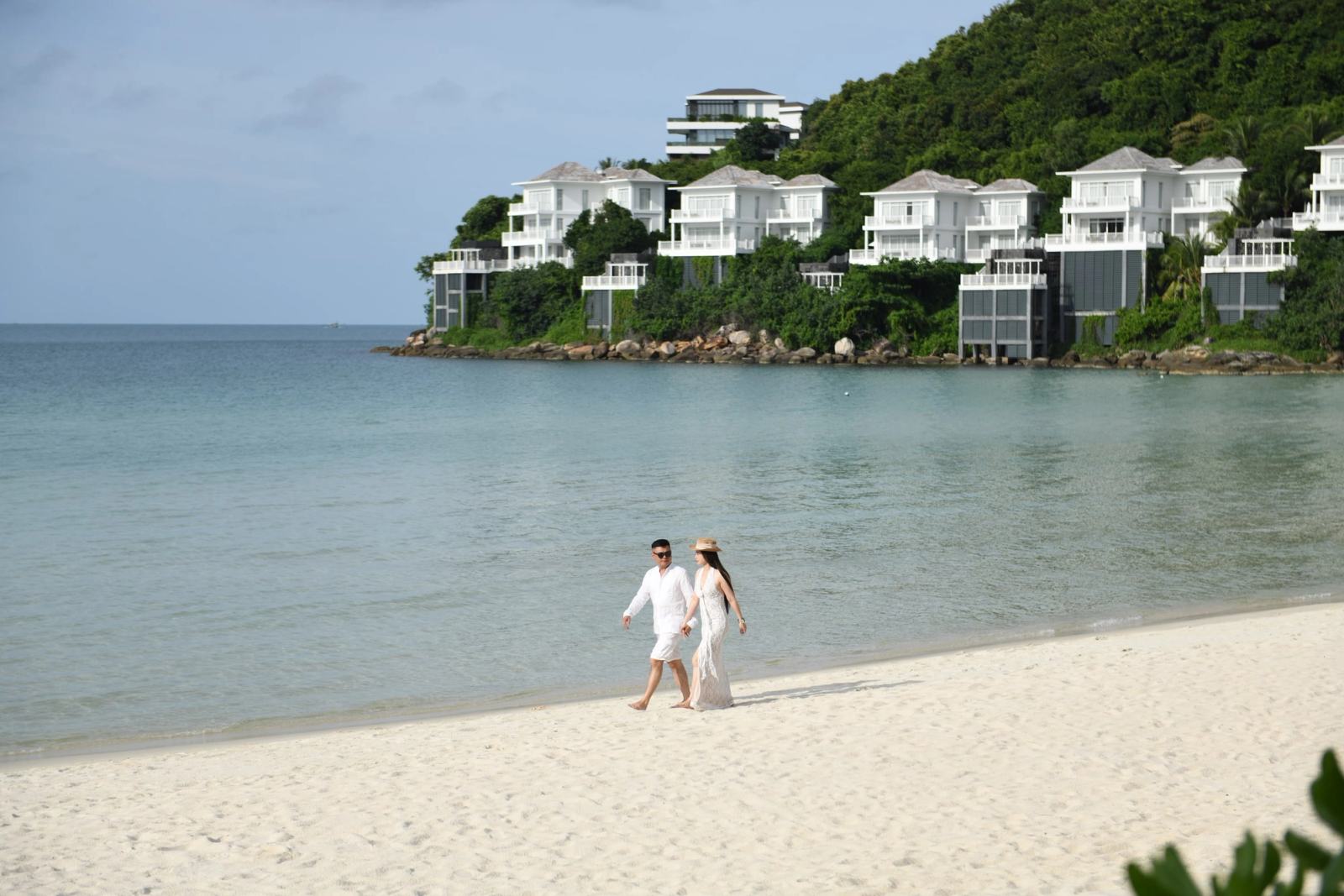 Couple walking along a sandy beach with white hillside villas across the bay.